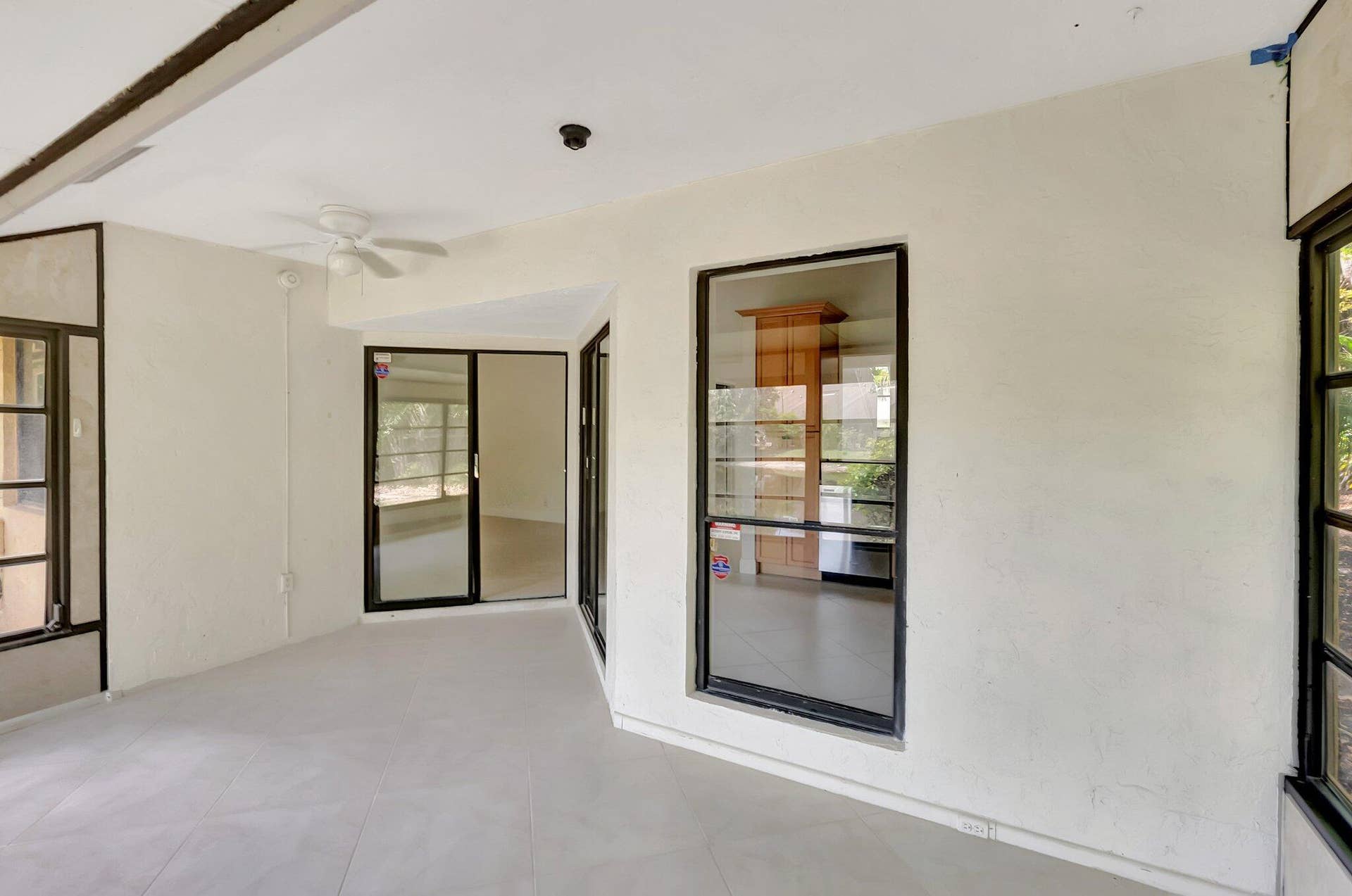 Spacious interior featuring large-format tile flooring, multiple floor-to-ceiling glass panel windows with black frames, sliding glass doors, and a ceiling fan