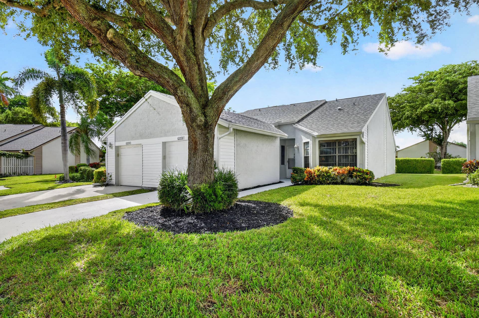 Single-story exterior featuring a light-colored siding facade, multi-gabled asphalt shingle roof, two-car garage, and mature landscaping with mulched tree rings