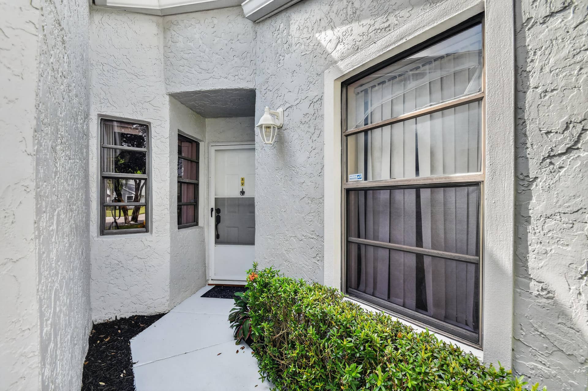 Textured stucco exterior featuring a white entry door, framed windows with metal casings, and exterior wall-mounted lighting