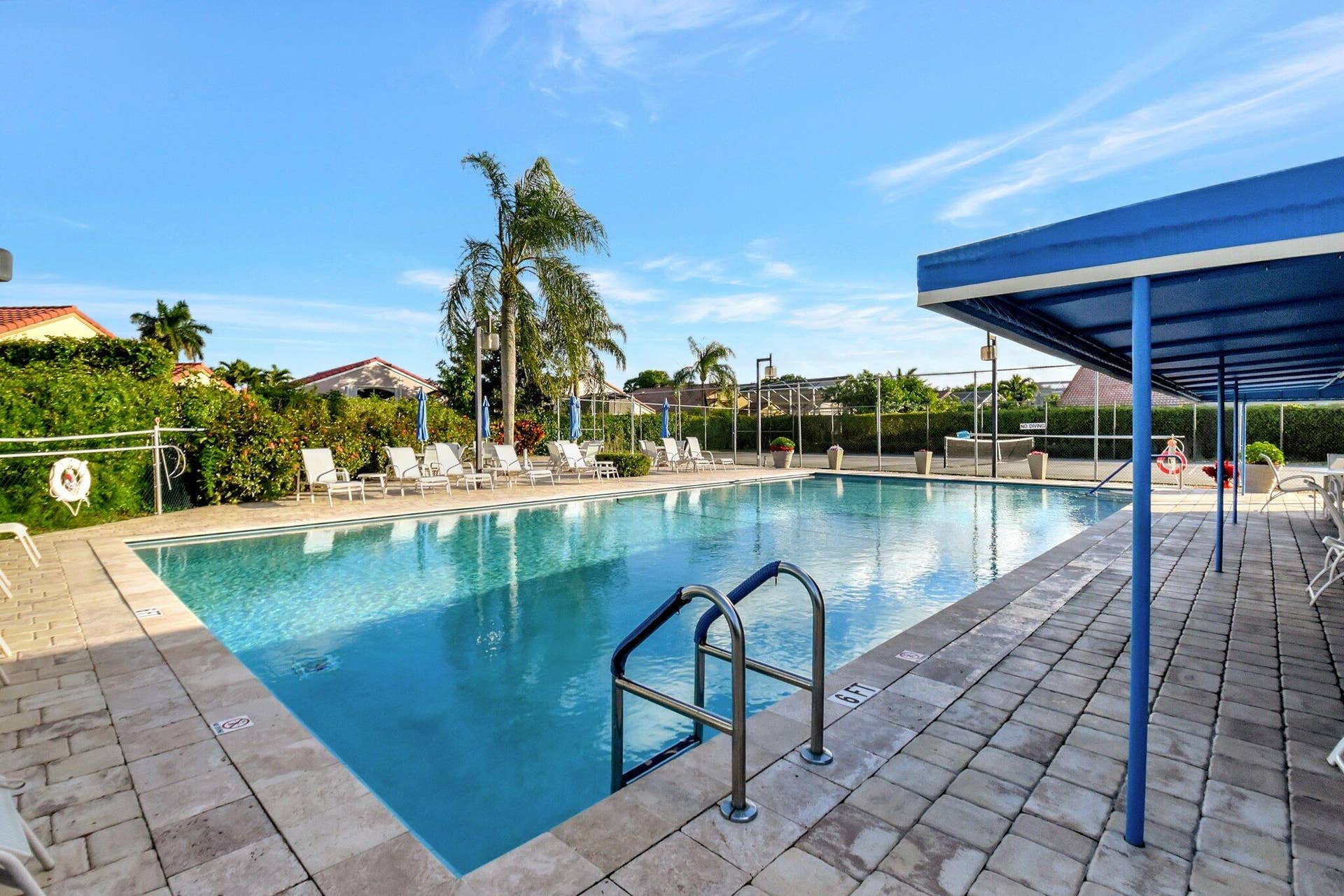 In-ground swimming pool with a stone paver deck, covered shade structure, metal handrails, and perimeter fencing
