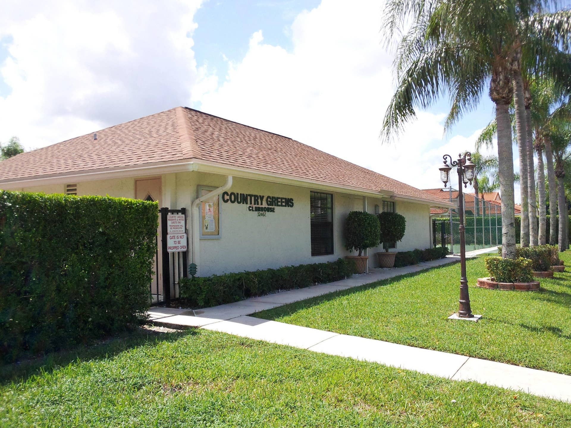 Community clubhouse exterior featuring a hip roof, stucco facade, manicured landscaping with mature palm trees, and an ornamental post-mounted light fixture