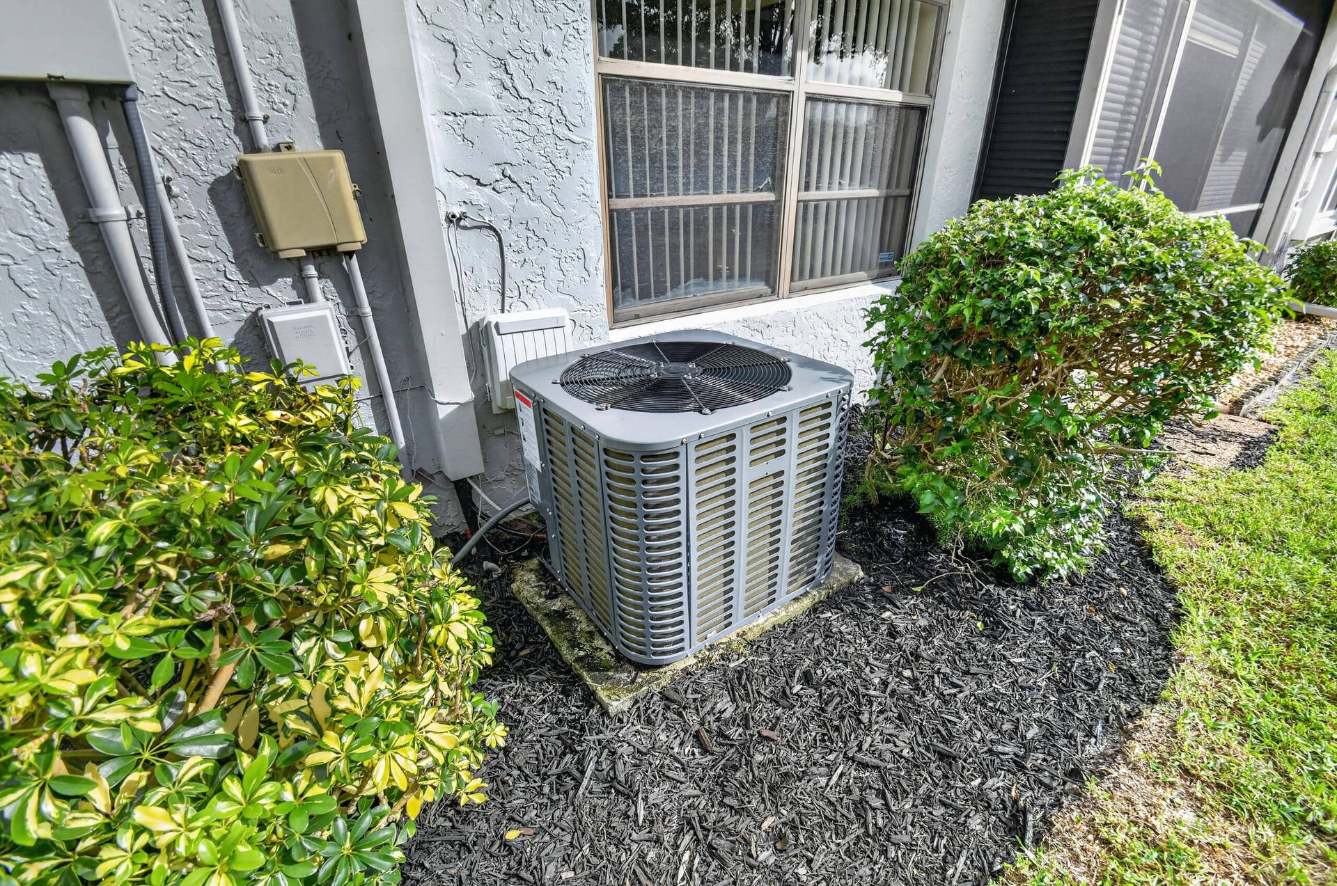 Exterior HVAC unit mounted on a concrete pad, surrounded by dark mulch and mature landscaping, featuring a stucco-textured exterior wall and grid-pattern window frame
