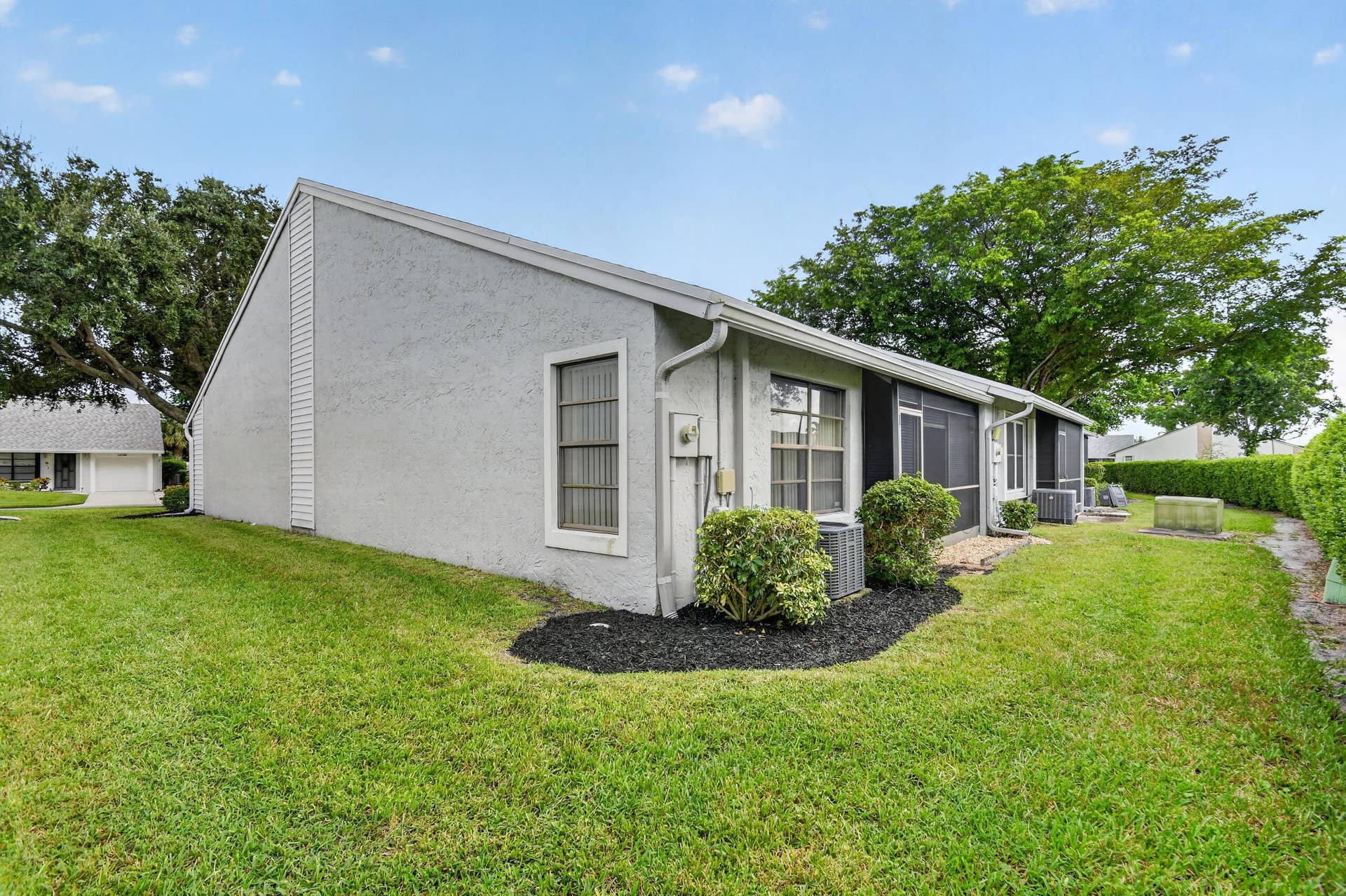Stucco exterior with a screened-in rear enclosure, integrated central air conditioning unit, and landscaped perimeter featuring mulched garden beds and mature trees