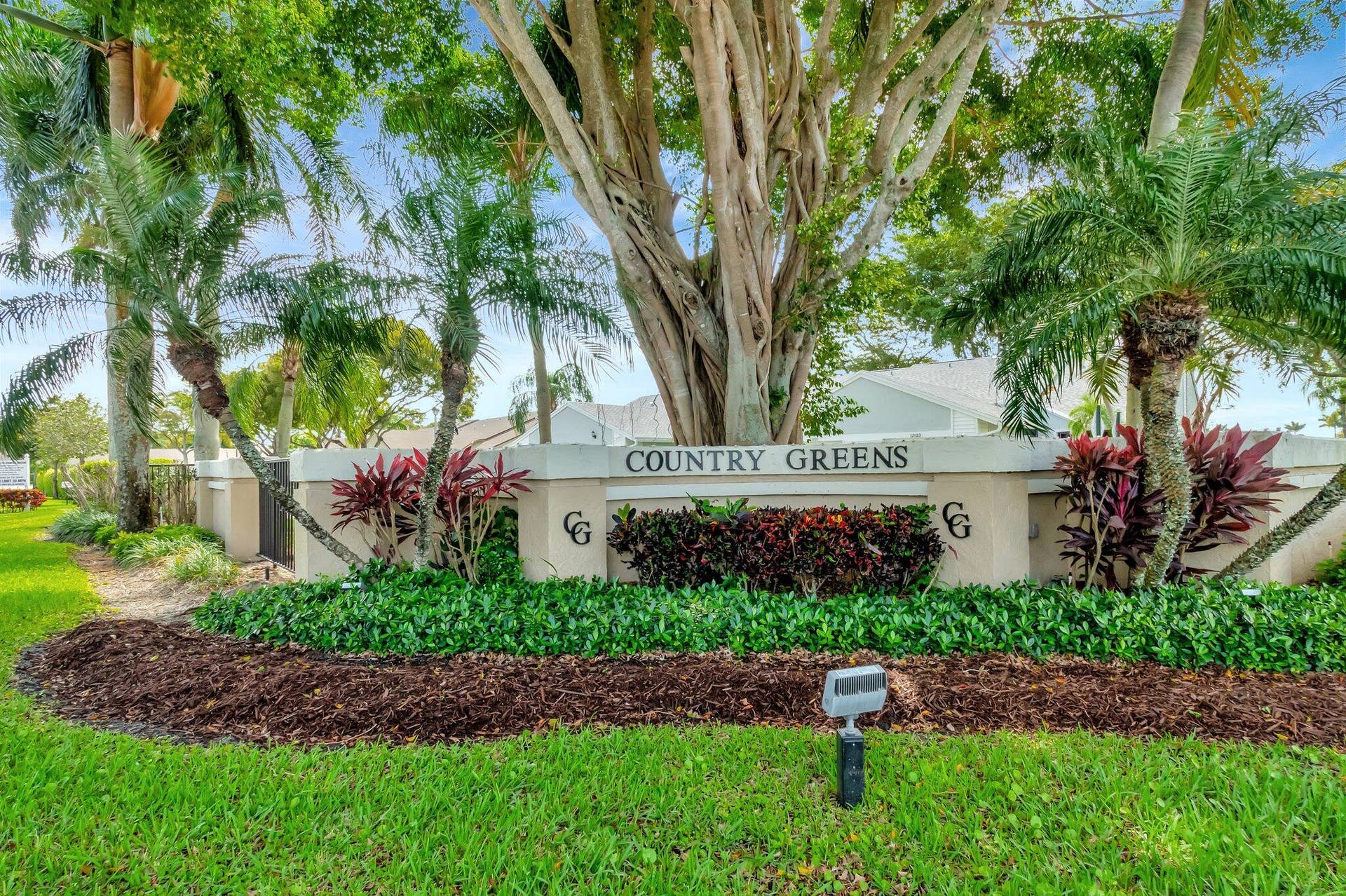 Stucco community monument sign featuring mature shade trees, established tropical landscaping, and mulched garden beds