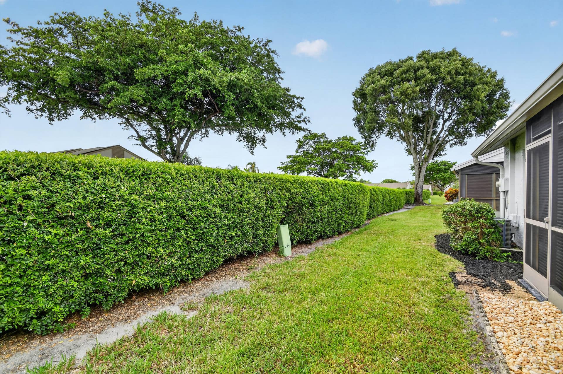 Rear exterior featuring a manicured lawn, established landscaping including a tall hedge row, mature trees, and a screened enclosure