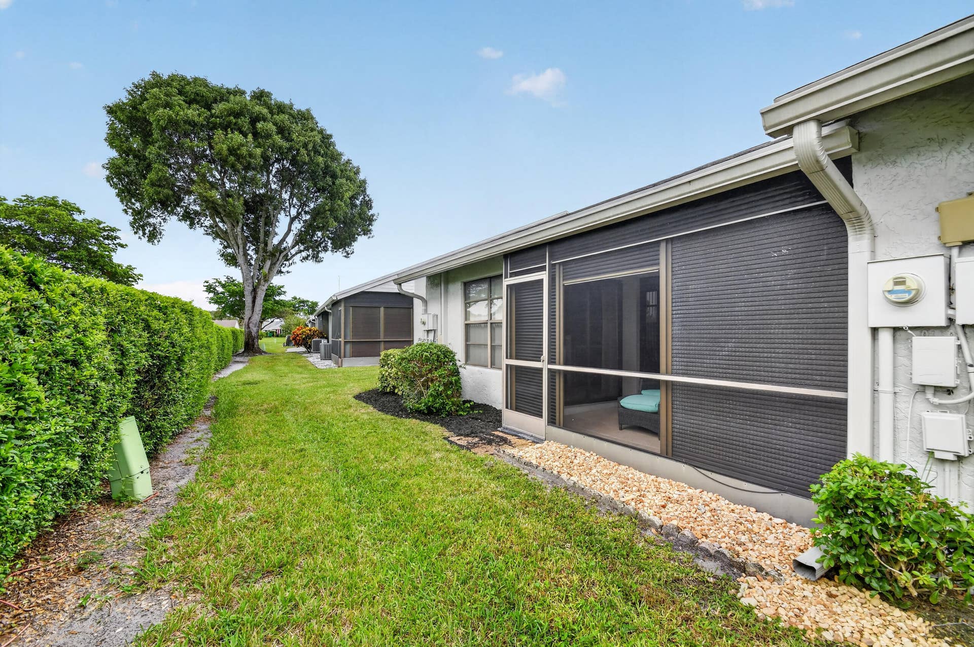 Stucco exterior featuring a screened-in patio, manicured hedge border, stone ground cover, and mature tree