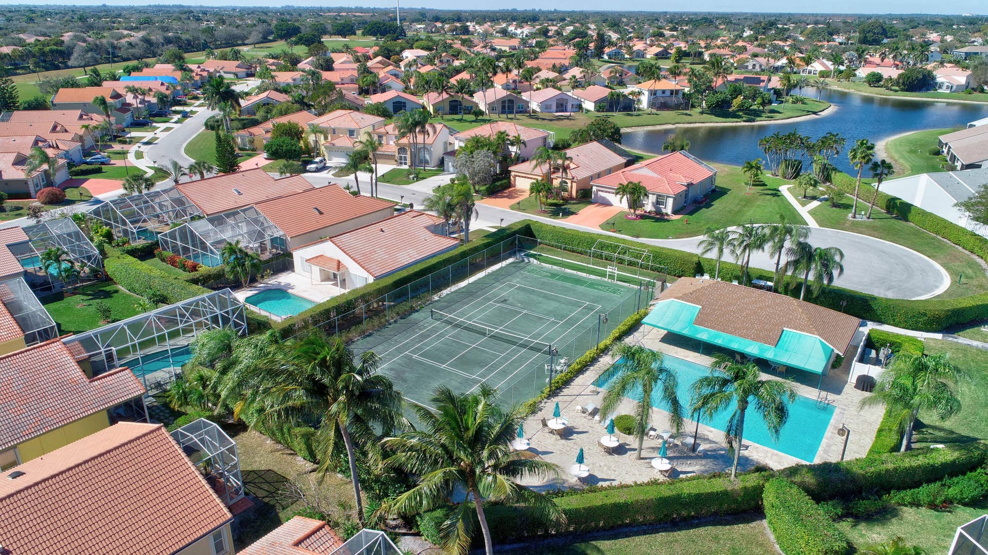 Aerial perspective of community amenities featuring a fenced tennis court, swimming pool with covered pavilion, and surrounding landscaping