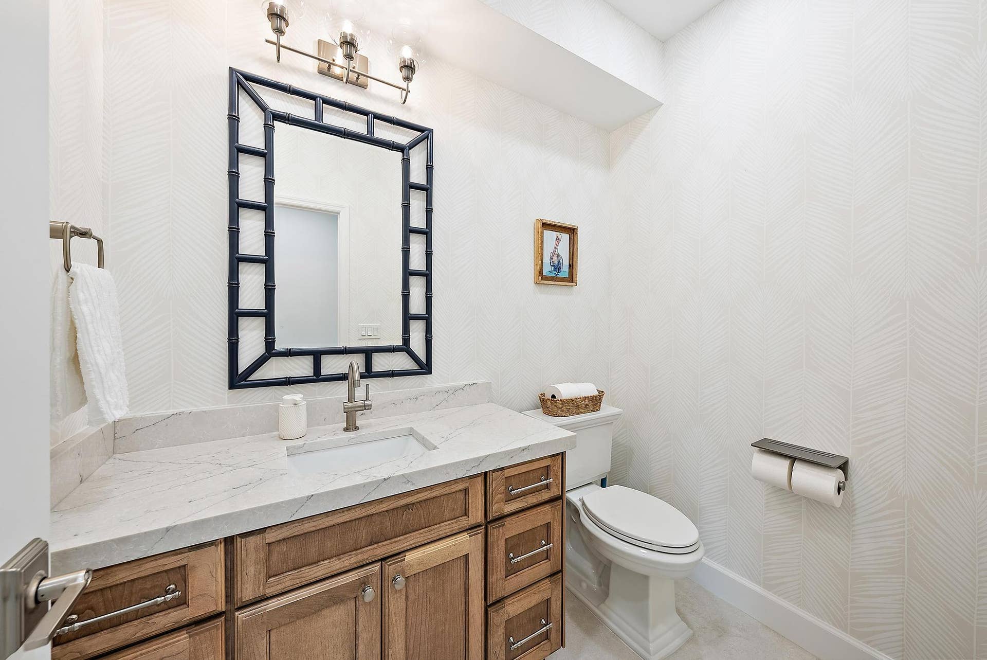 Natural-finish wood cabinetry, stone-surface countertop with an integrated sink, polished metal faucet, decorative mirror frame, and textured wallpaper