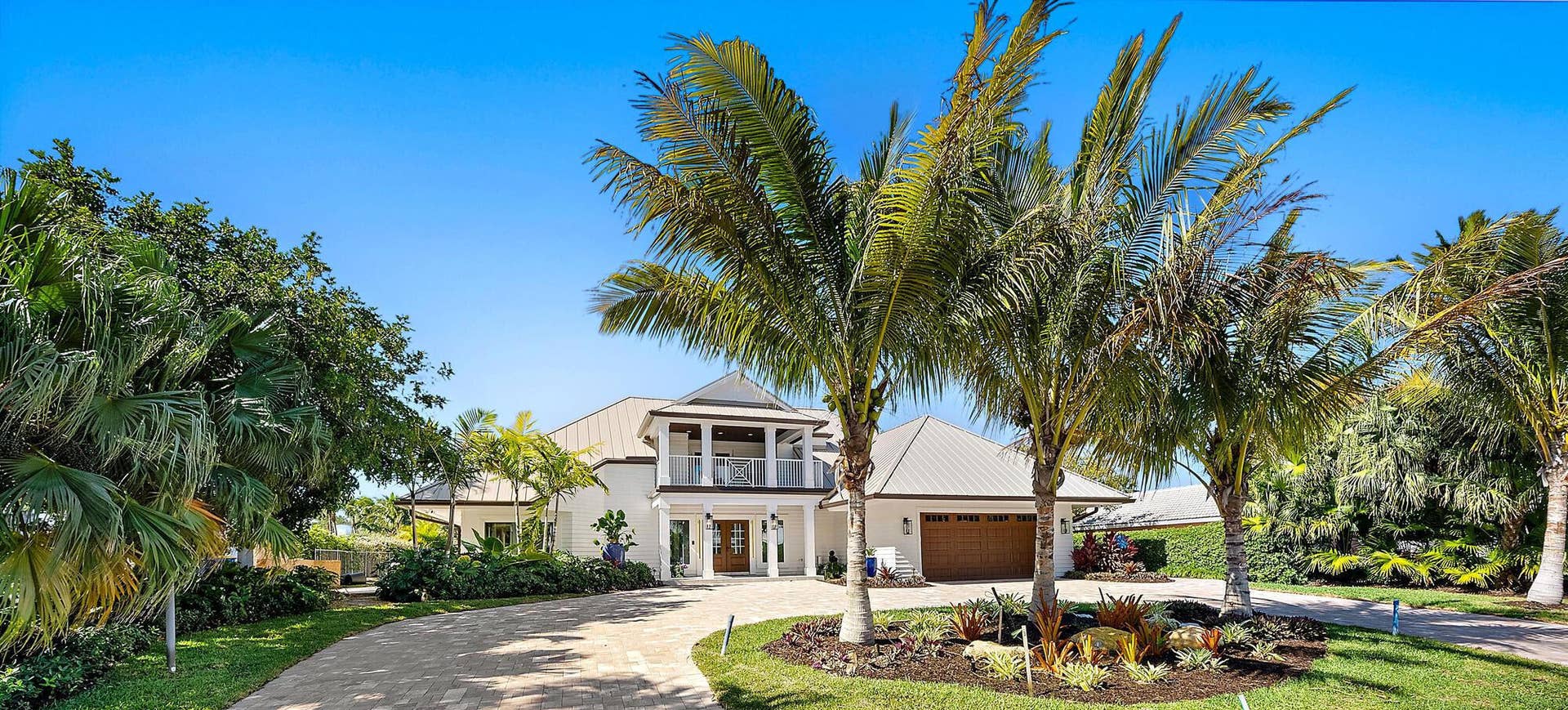 Two-story exterior featuring a metal roof, balcony with decorative railings, multi-car garage, and a paved circular driveway surrounded by mature palm trees and landscaped garden beds