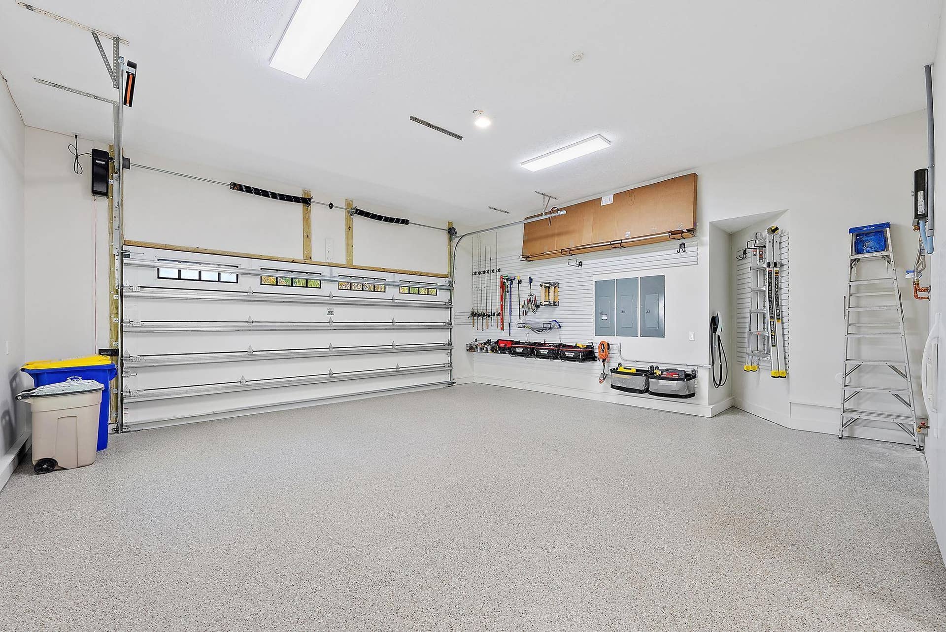 Spacious garage featuring an epoxy-coated floor, integrated wall-mounted slat board storage system, overhead fluorescent lighting, and a multi-panel sectional door