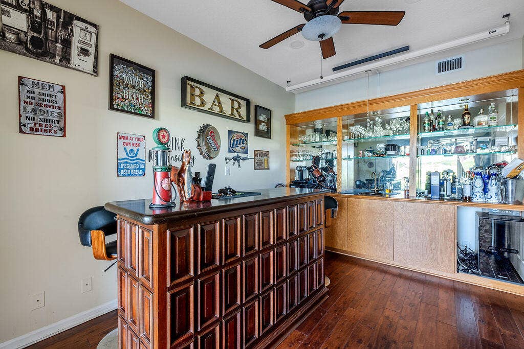 1233 Avondale Lane, West Palm Beach, FL 33409 Built-in wood wet bar featuring recessed panel cabinetry, integrated shelving with glass accents, a dark stone countertop, and a ceiling-mounted fan
