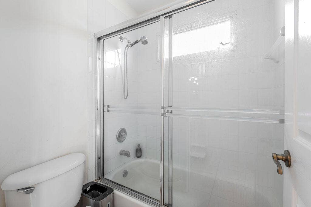 1233 Avondale Lane, West Palm Beach, FL 33409 Full-size bathtub with white tile surround, sliding glass shower doors, adjustable handheld shower fixture, and recessed window
