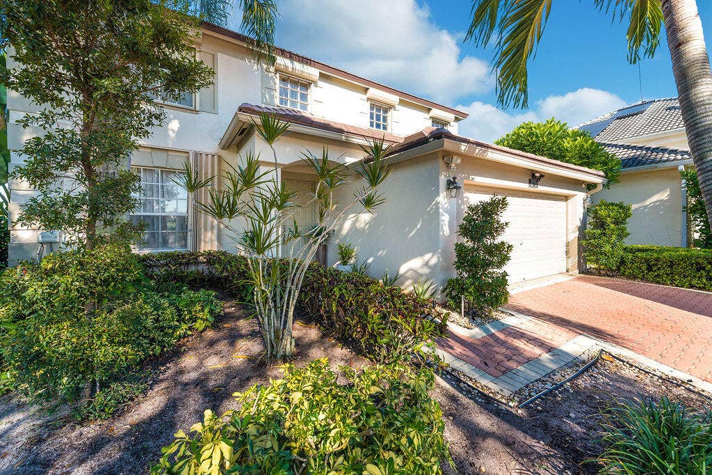 1233 Avondale Lane, West Palm Beach, FL 33409 Two-story stucco exterior featuring a covered entryway, attached garage, and paver driveway surrounded by mature landscaping