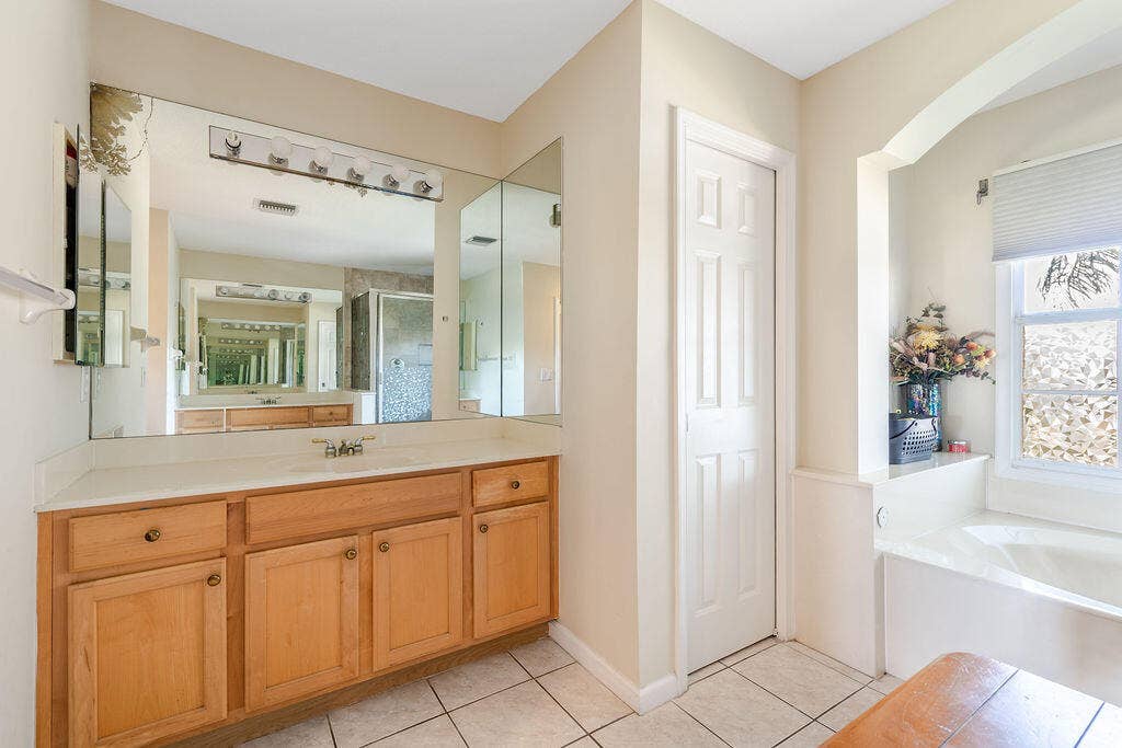 1233 Avondale Lane, West Palm Beach, FL 33409 Bathroom featuring a wood-finish vanity with a light-toned countertop, a multi-panel wall mirror, ceramic tile flooring, and a recessed soaking tub within a dedicated alcove