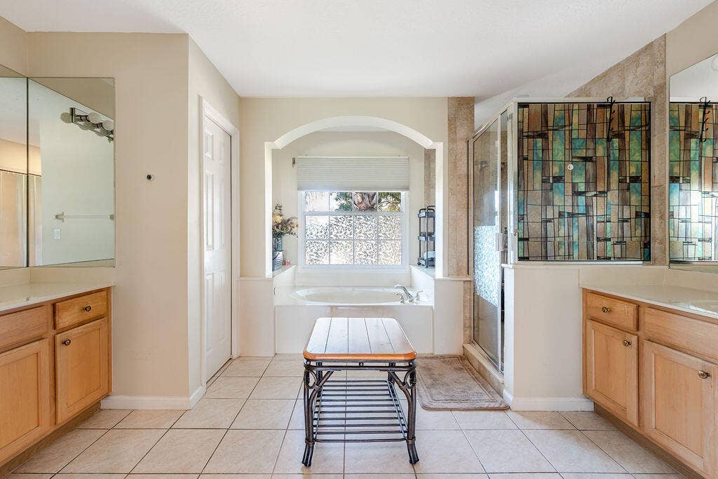 1233 Avondale Lane, West Palm Beach, FL 33409 Bathroom featuring dual wood-finish vanities, a tiled soaking tub set within an arched alcove, a glass-enclosed shower with geometric patterned tile accents, and neutral floor tiling