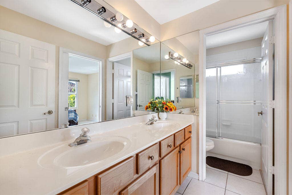 1233 Avondale Lane, West Palm Beach, FL 33409 Dual-sink vanity with a solid-surface countertop, full-width wall mirror, wood-finish cabinetry, and a shower-tub combination with sliding glass doors
