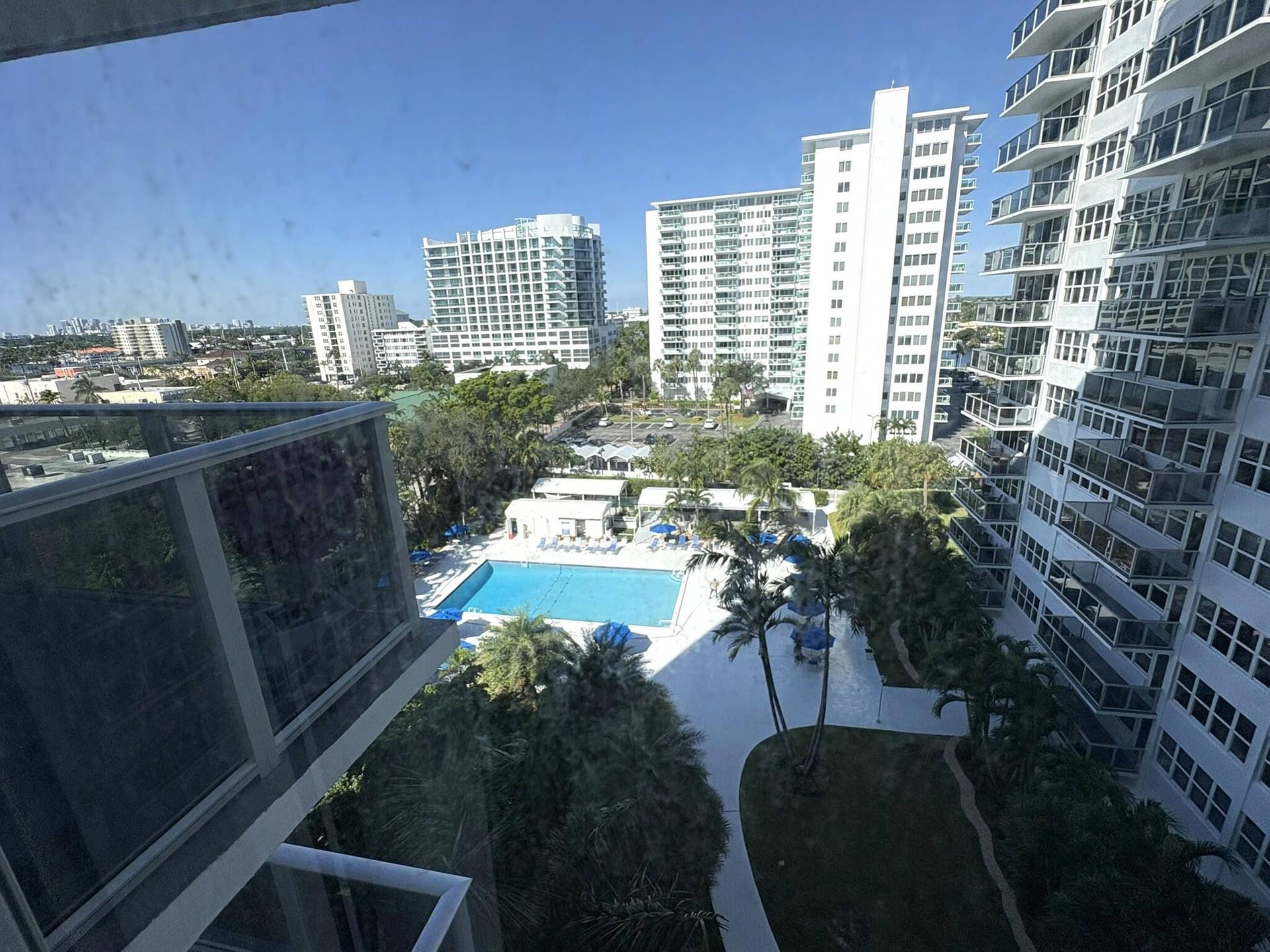 Balcony featuring glass railings, overlooking a swimming pool, sun deck, paved pathways, and mature palm trees