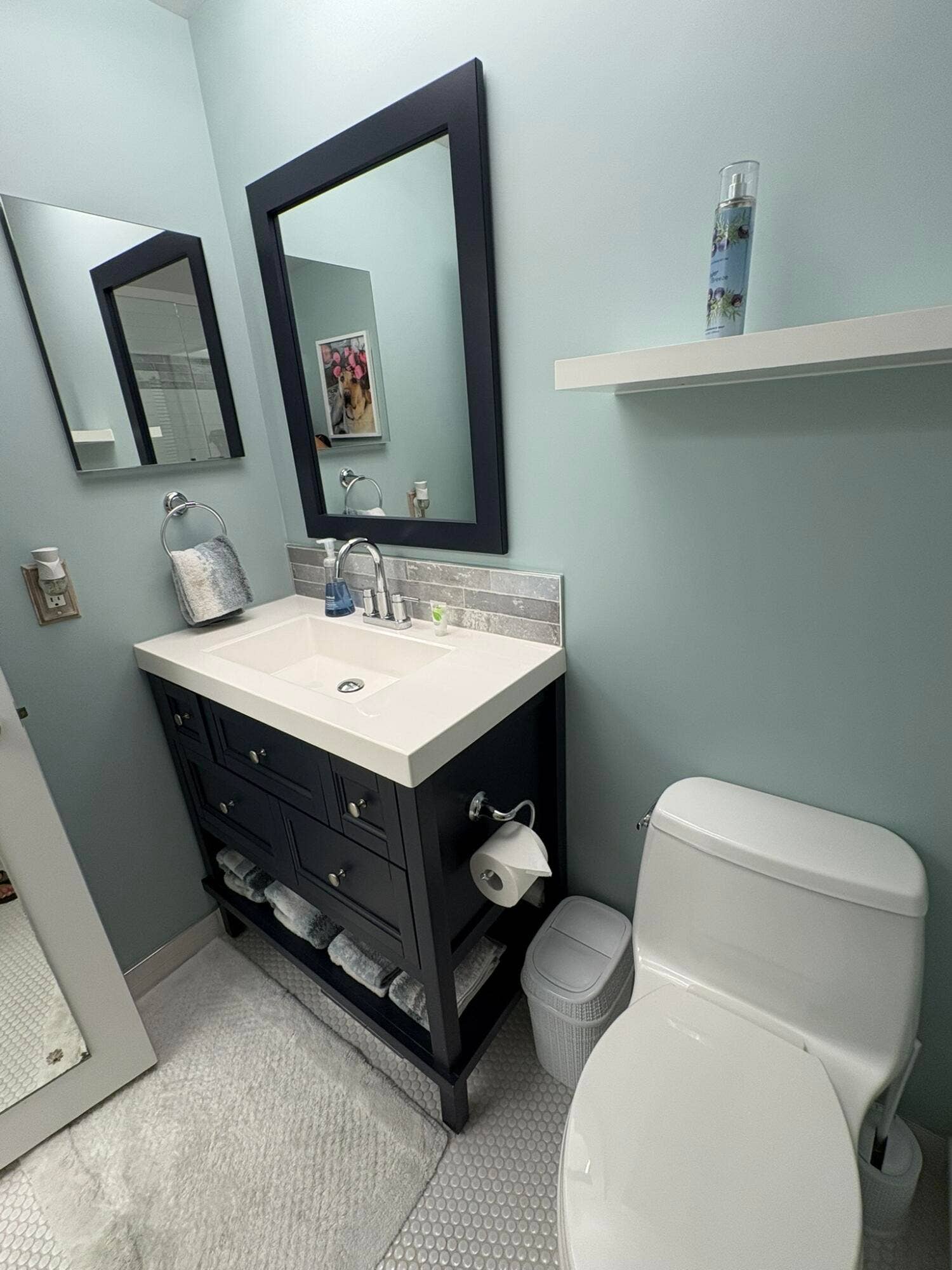 Dark-finish vanity with an integrated rectangular sink and stone-look backsplash, paired with a matching framed mirror, white wall-mounted shelving, and hexagonal tile flooring