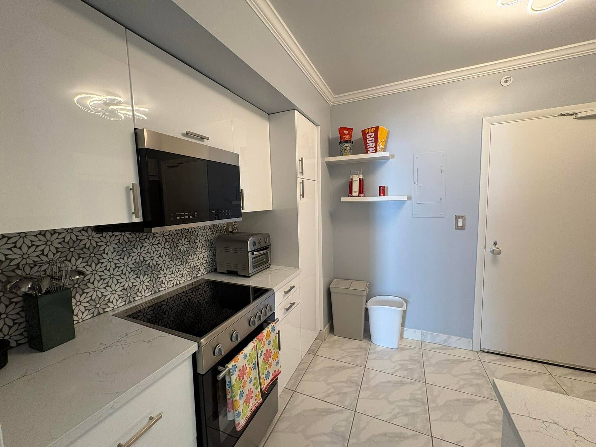 High-gloss white cabinetry, stone-finish countertops, floral-patterned tile backsplash, stainless steel microwave, and large-format tile flooring