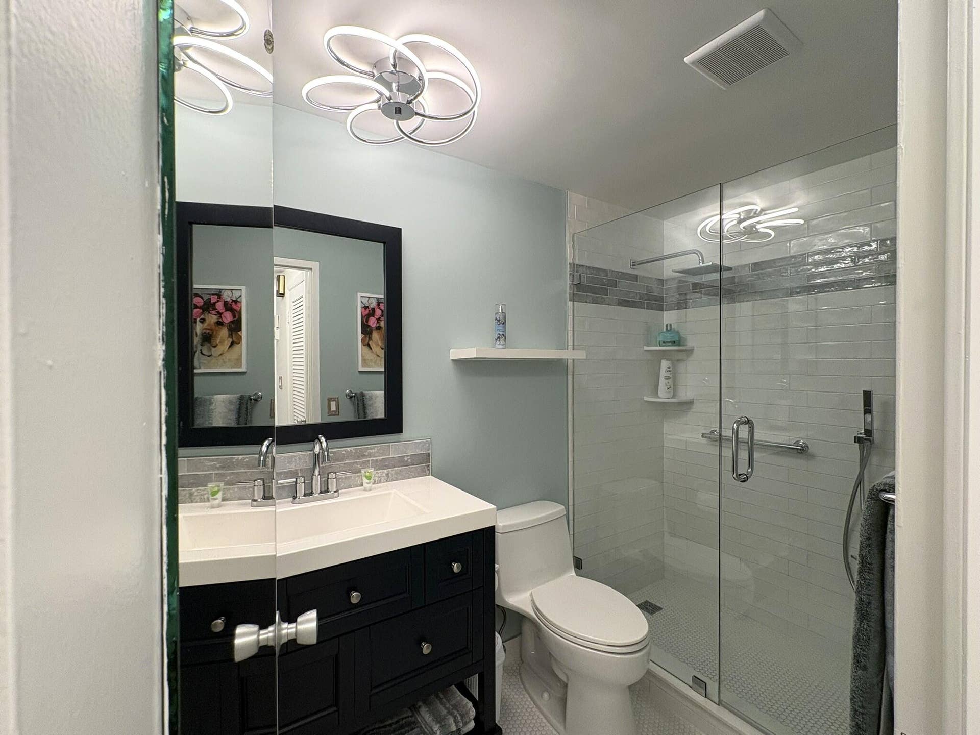 Bathroom featuring a dark wood-finish vanity with an integrated basin, contemporary circular LED light fixture, glass-enclosed shower with subway tile surround, decorative tile inlay, and hexagonal floor tile