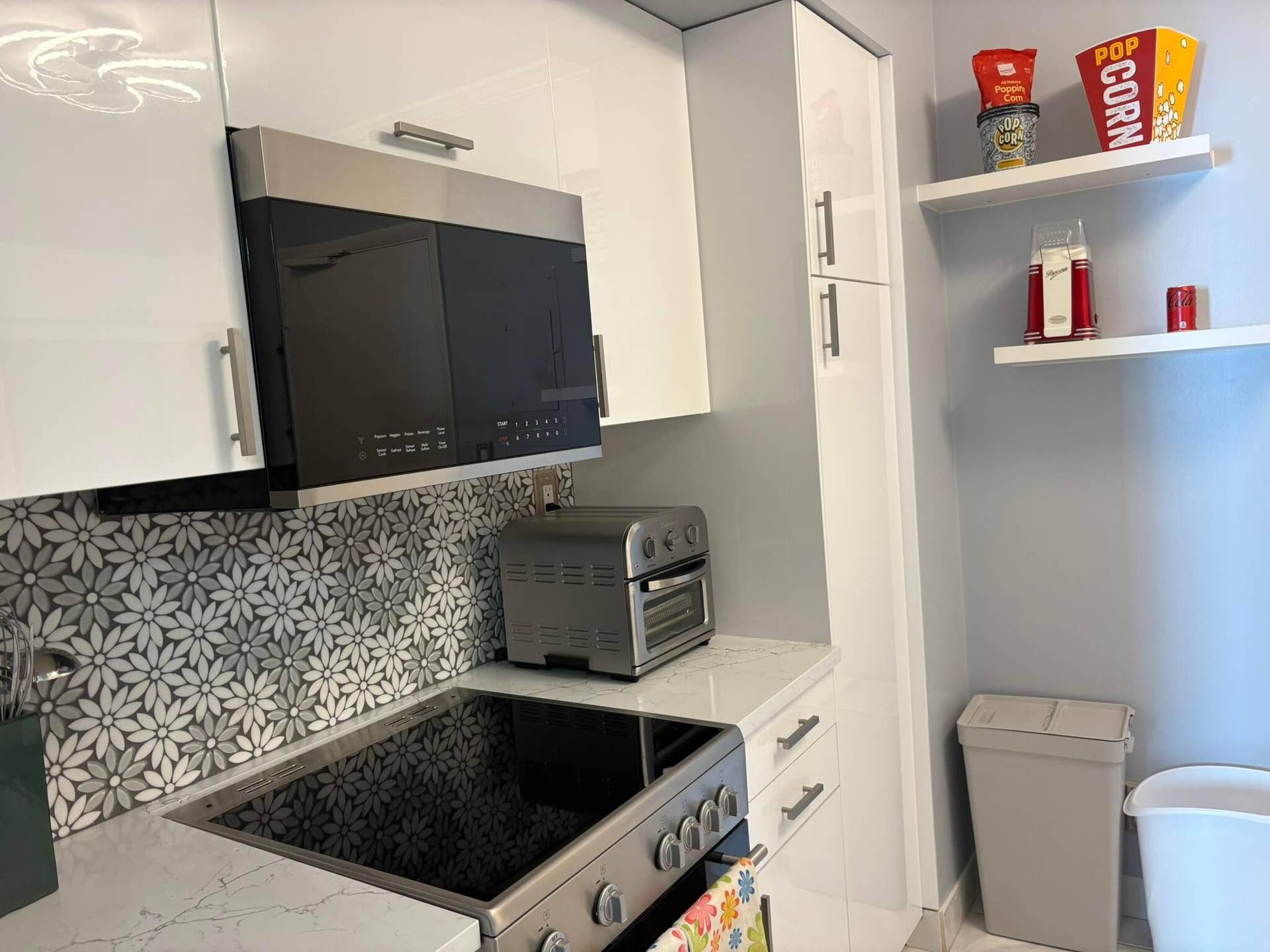 Kitchen featuring high-gloss white cabinetry, stone-finish countertops, stainless steel microwave, floral-pattern tile backsplash, and built-in floating shelves