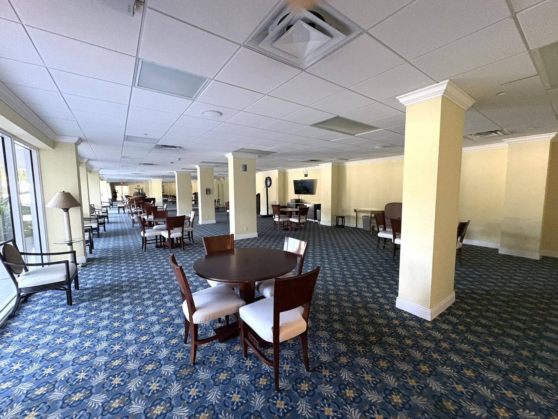 Open-concept interior featuring drop ceiling tiles, structural columns with crown molding, patterned carpet flooring, and floor-to-ceiling windows