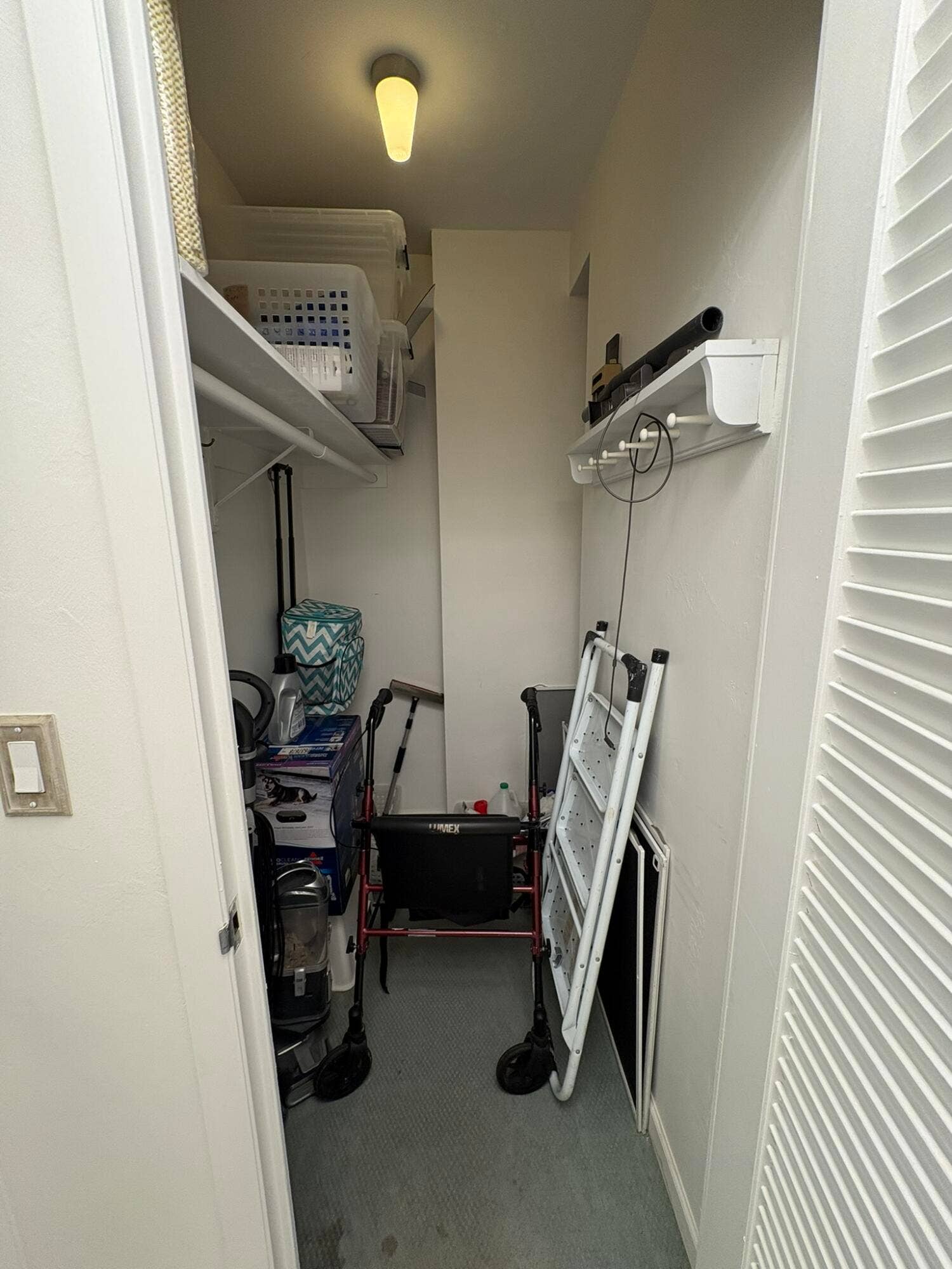 Storage closet featuring a ceiling-mounted light fixture, upper shelving, and a wall-mounted hook rack