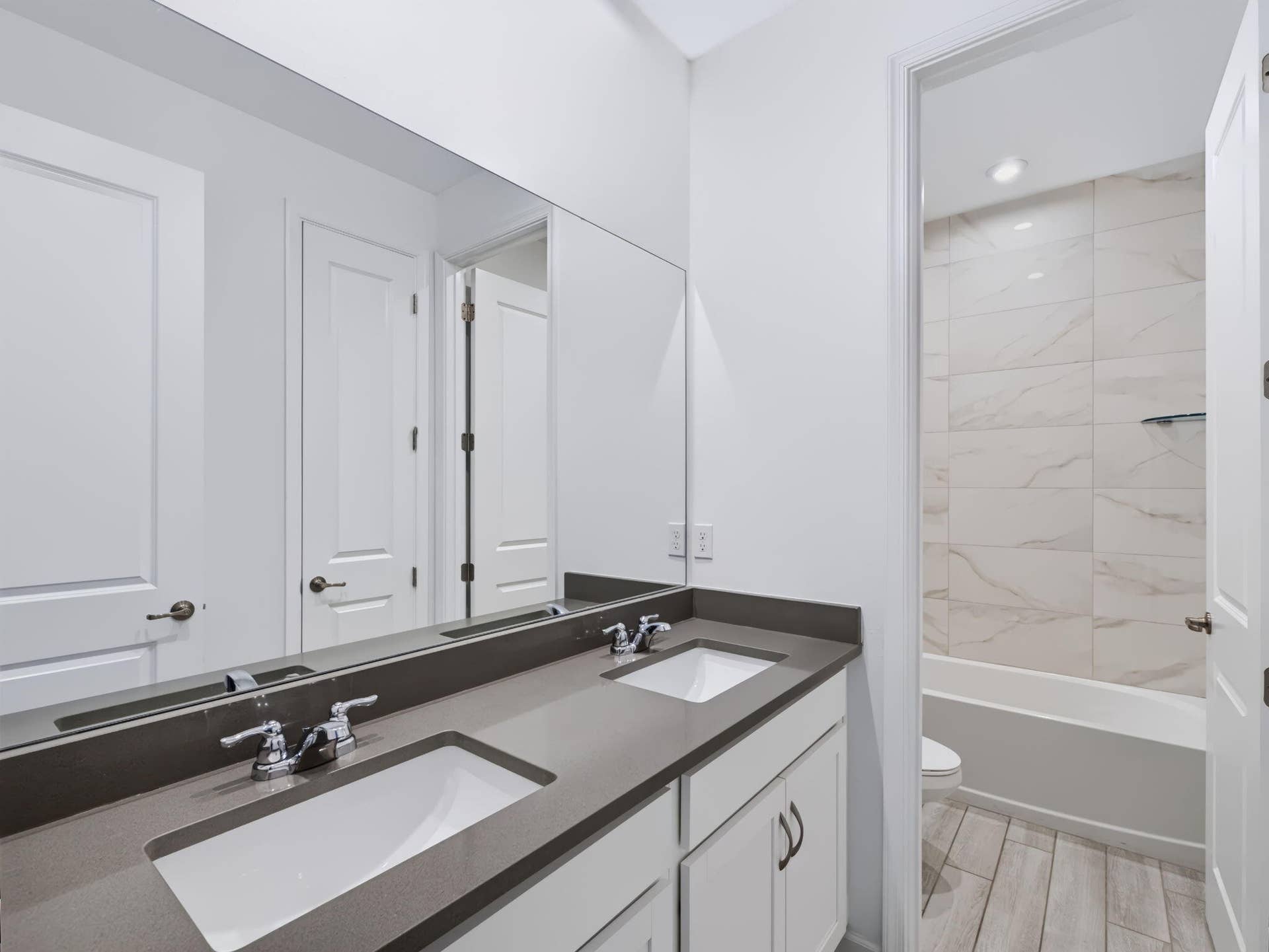 12688 SW Eleanor Drive, Port St Lucie, FL 34987 Dual-sink vanity with a solid-surface countertop, full-width wall mirror, wood-finish flooring, and a tub-shower combination with floor-to-ceiling tile surround