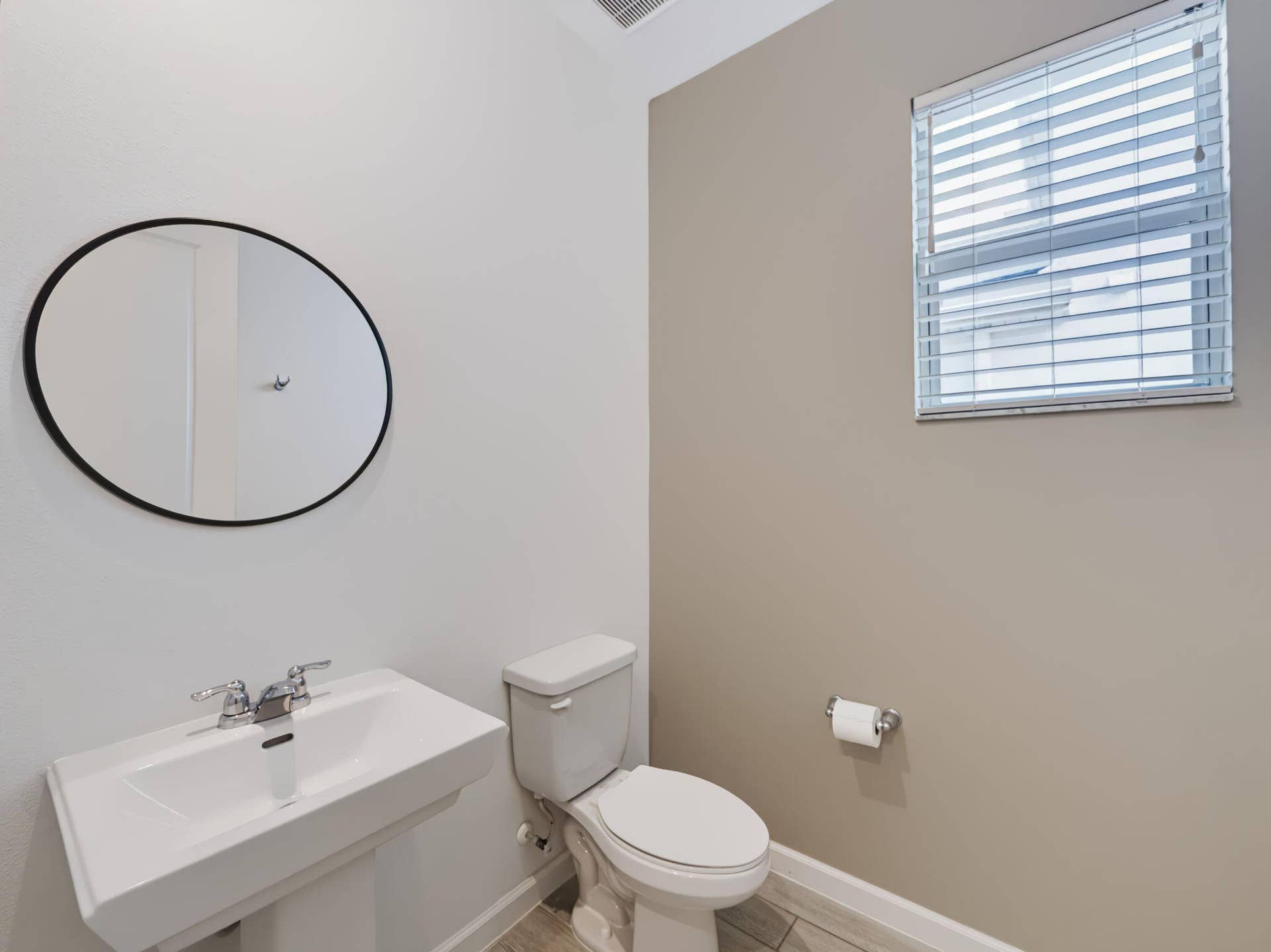 12688 SW Eleanor Drive, Port St Lucie, FL 34987 Wall-mounted oval mirror with a black frame, white pedestal sink featuring dual-handle chrome hardware, and horizontal blind window treatment