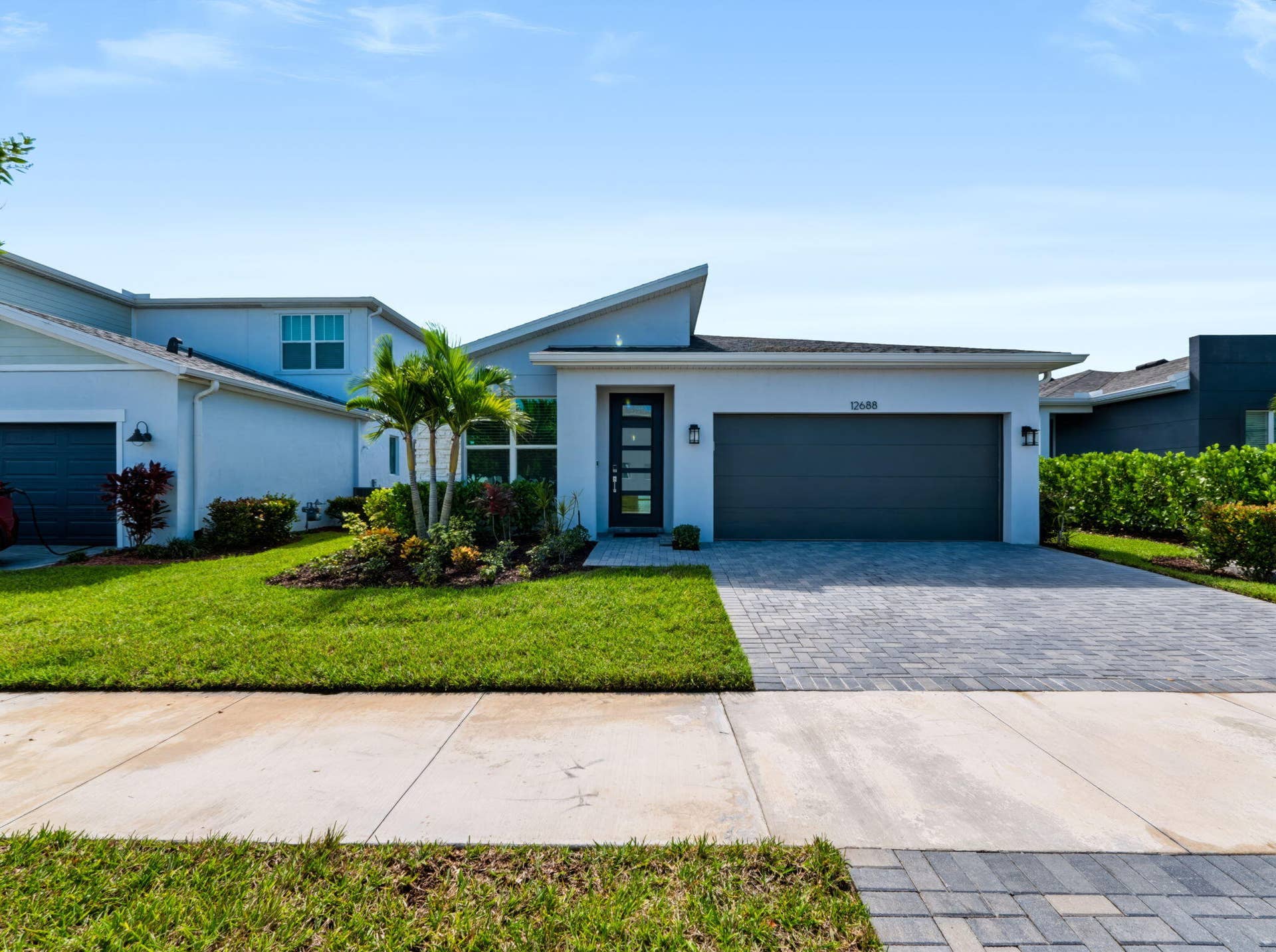 12688 SW Eleanor Drive, Port St Lucie, FL 34987 Modern exterior featuring a geometric roofline, paver driveway, two-car garage, glass-panel front door, and manicured landscaping