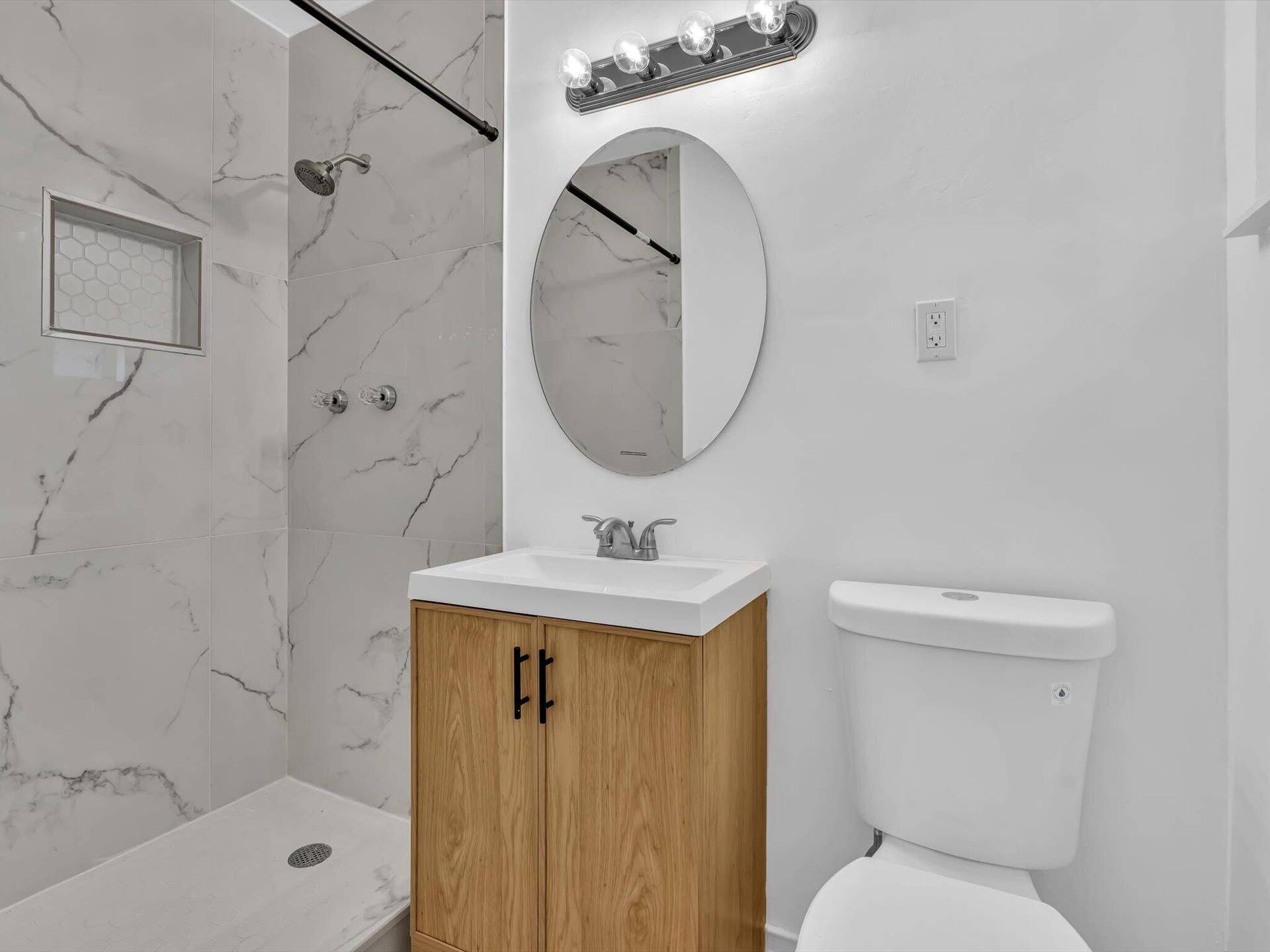 132 SE 2ND Court, Dania Beach, Dania Beach, FL 33004 Bathroom featuring large-format veined tile shower surround with integrated recessed niche, wood-finish vanity with rectangular basin, oval wall-mounted mirror, and multi-light overhead vanity fixture