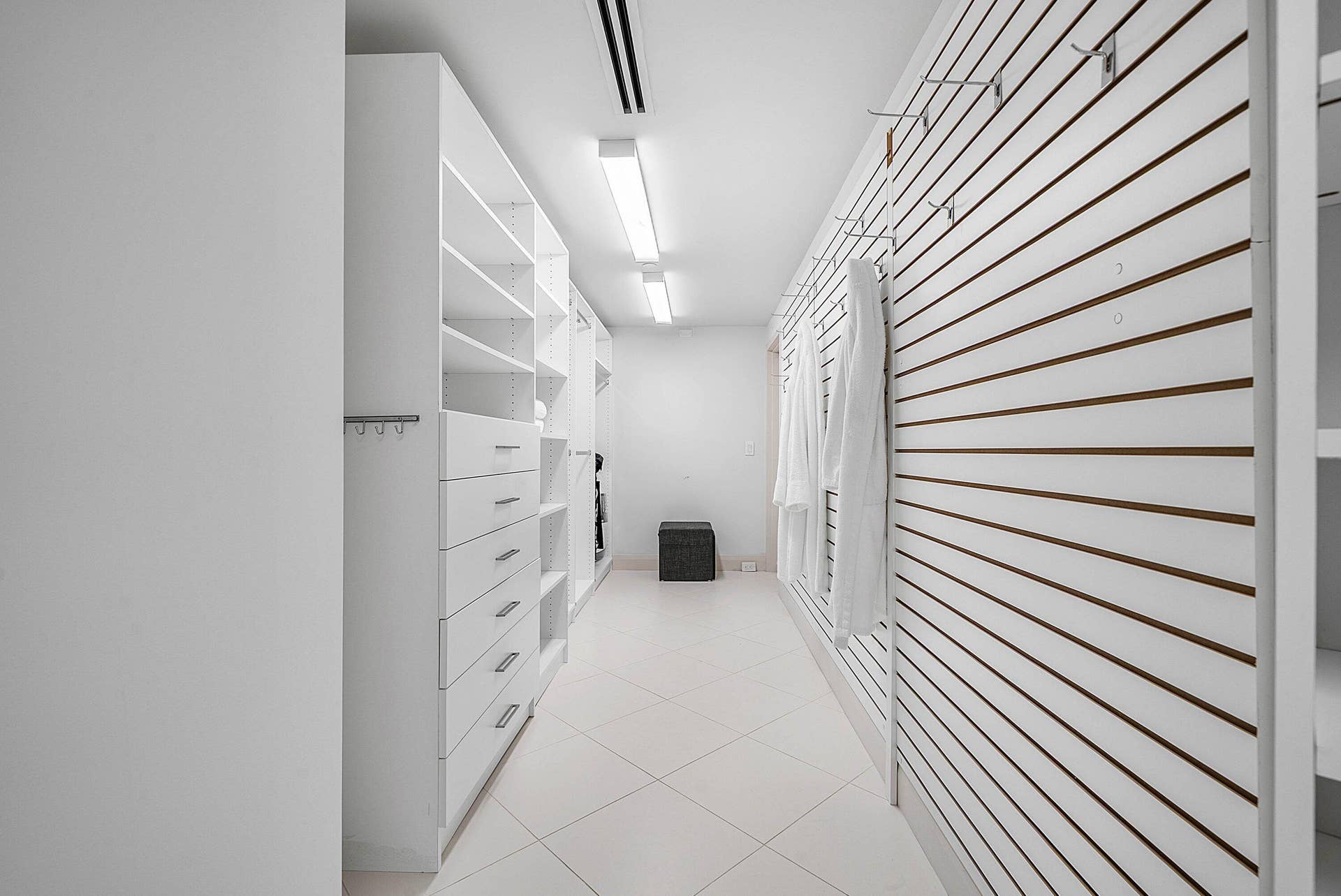 Walk-in closet featuring white built-in cabinetry with integrated drawers, slat-wall organizational system with adjustable hooks, tile flooring, and linear flush-mount lighting