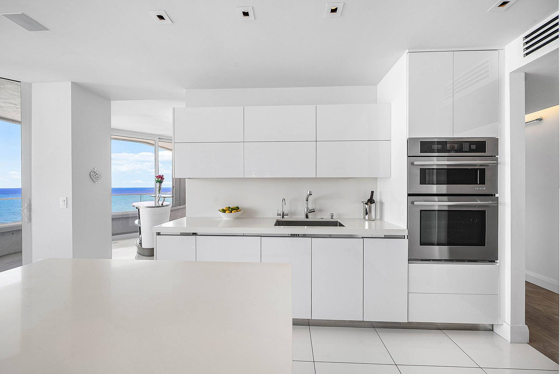 Contemporary kitchen featuring high-gloss white cabinetry, integrated stainless steel double wall ovens, solid-surface countertops, recessed ceiling lighting, and large-format tile flooring