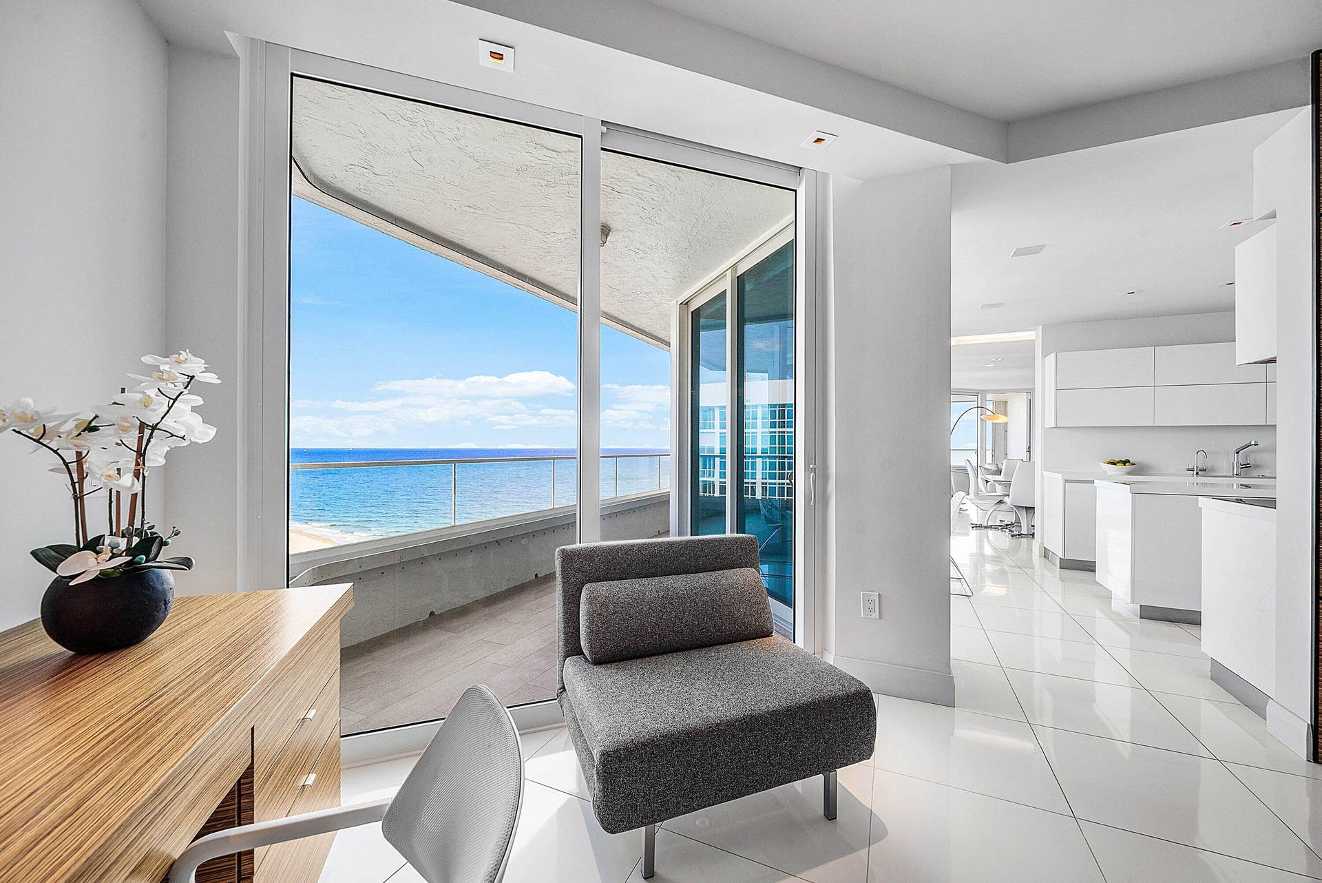 Floor-to-ceiling glass sliding doors opening to an expansive balcony with ocean scenery, white tile flooring, and sleek flat-panel cabinetry