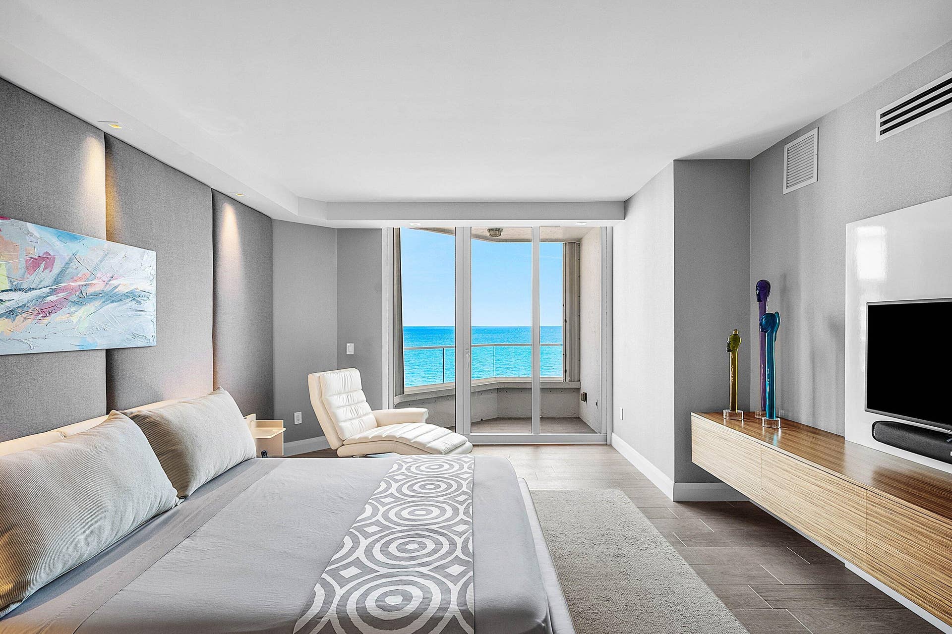 Floor-to-ceiling glass sliding doors opening to a balcony, wall-to-wall upholstered headboard paneling, wall-mounted wood-finish media console, and large-format tile flooring