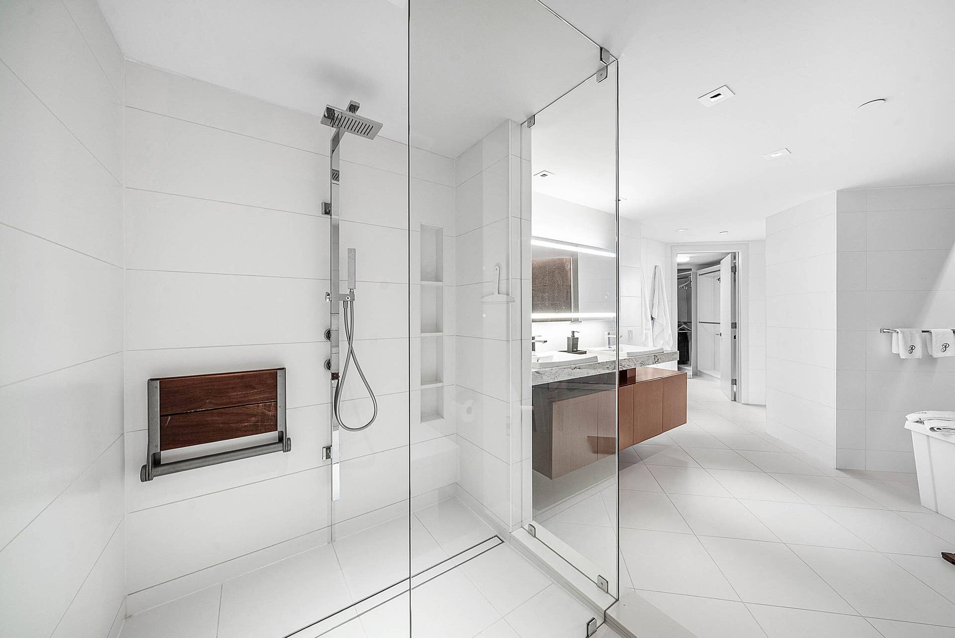Walk-in shower with floor-to-ceiling tile, frameless glass enclosure, wall-mounted shower panel with handheld sprayer, built-in shelving, and integrated wooden fold-down seat