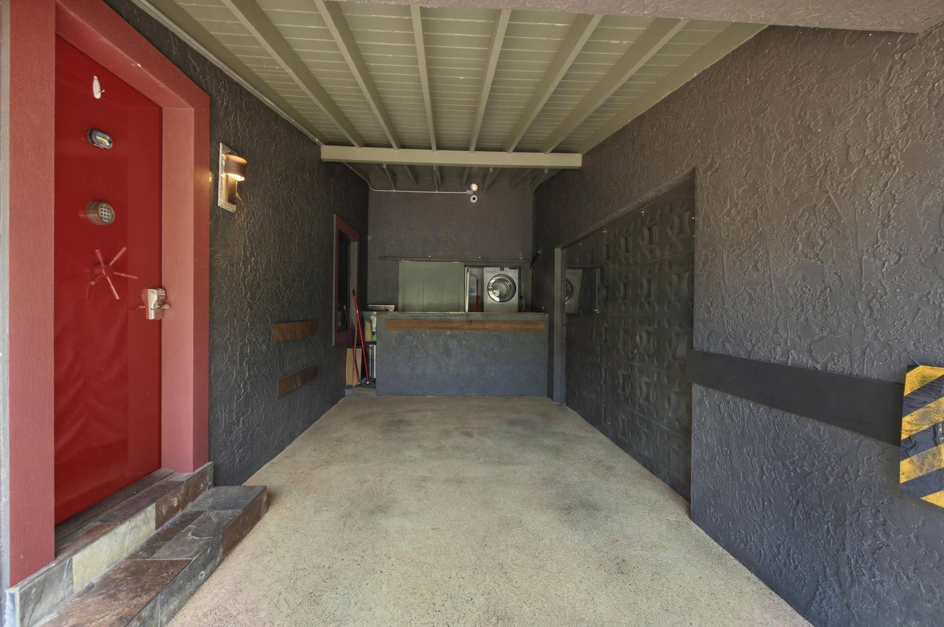 1110 SW 18TH Court, Fort Lauderdale, FL 33315 Covered carport featuring a vibrant red entry door, textured dark wall cladding, concrete flooring, and an elevated storage platform
