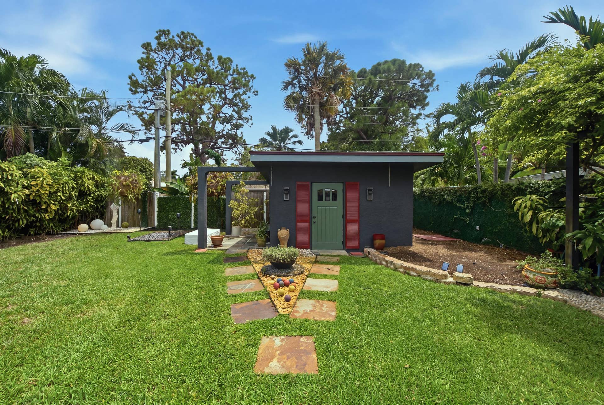 1110 SW 18TH Court, Fort Lauderdale, FL 33315 Detached structure featuring a masonry exterior, decorative red shutters, a green paneled entry door, a stone-paved walkway, and mature landscaping