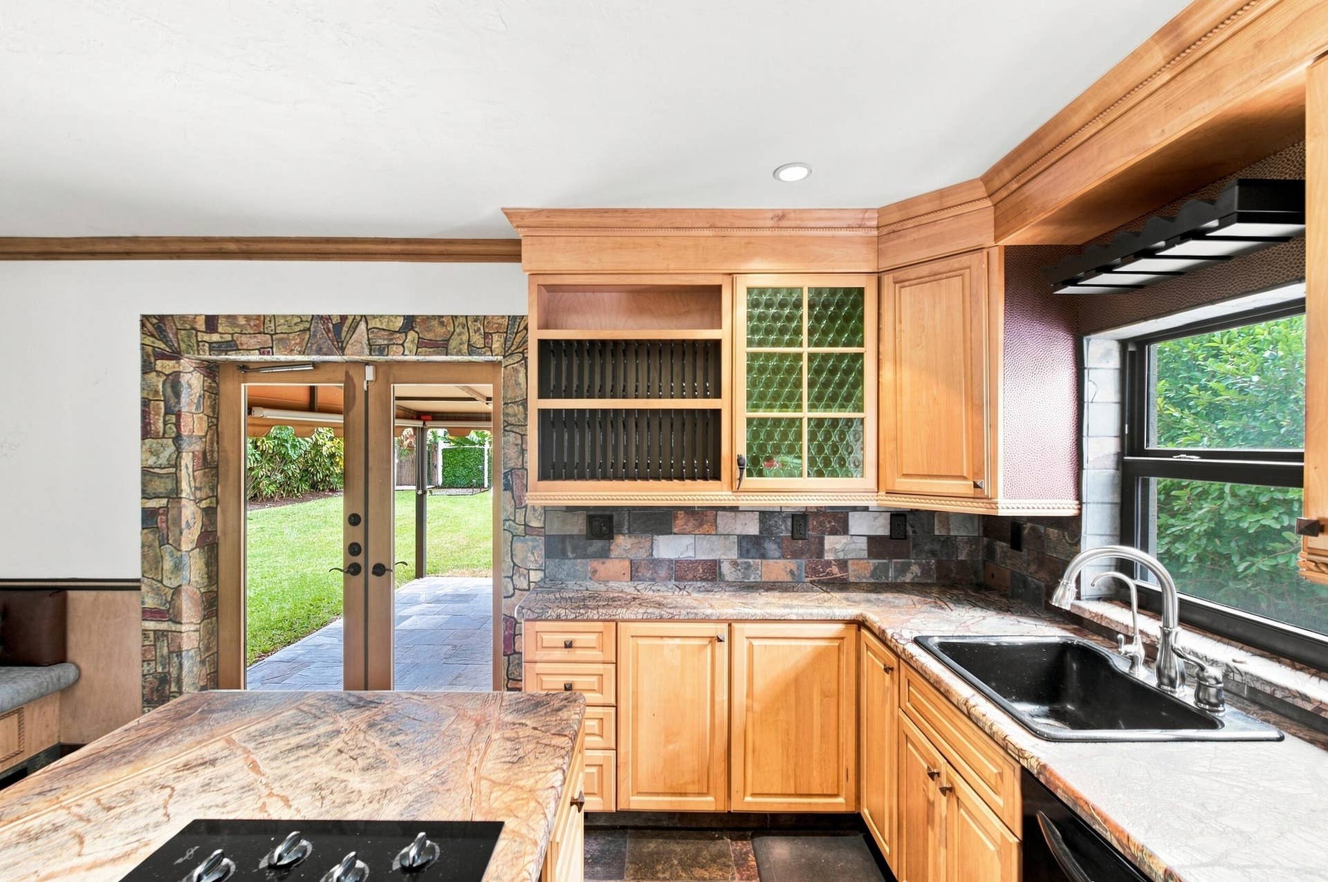 1110 SW 18TH Court, Fort Lauderdale, FL 33315 Kitchen featuring custom natural wood cabinetry, stone tile backsplash, integrated stone-topped island with stovetop, drop-in sink, and double glass doors encased in stone masonry