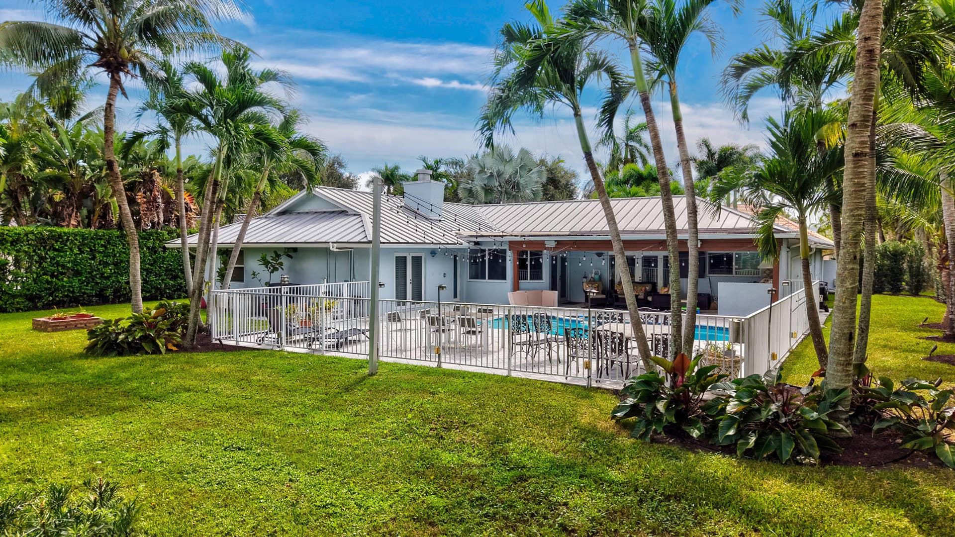 2919 Banyan Blvd Circle NW, Boca Raton, FL 33431 Rear exterior featuring a metal roof, a fenced-in swimming pool, a covered patio area, and mature palm trees within a landscaped yard