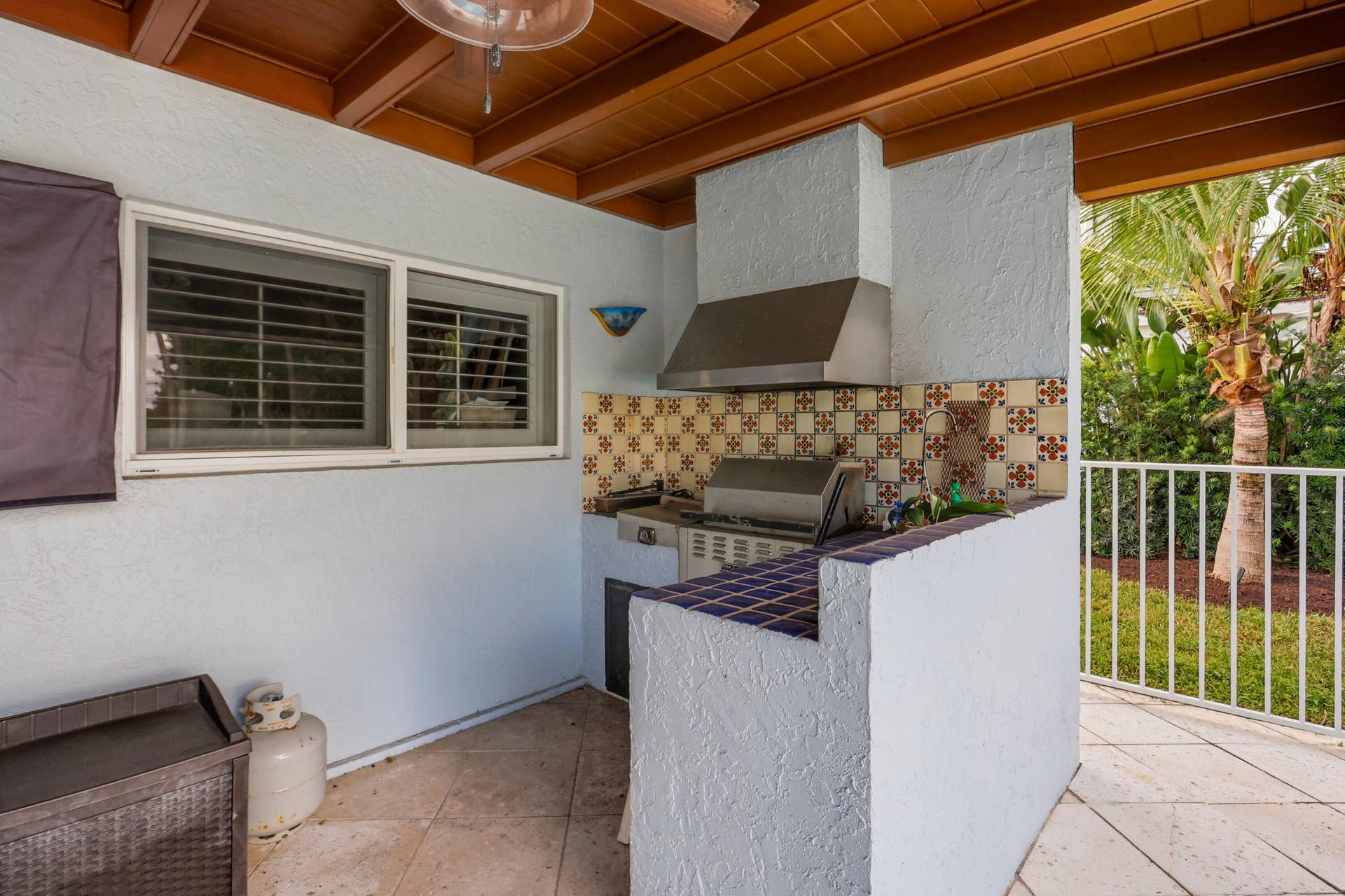2919 Banyan Blvd Circle NW, Boca Raton, FL 33431 Covered outdoor kitchen featuring a built-in stainless steel grill, range hood, tiled backsplash, and tile countertop with integrated sink, situated under a wood-beam ceiling