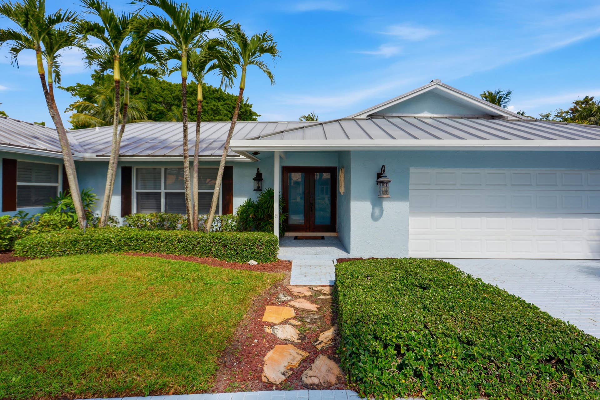 2919 Banyan Blvd Circle NW, Boca Raton, FL 33431 Single-story residence featuring a standing seam metal roof, stone-paved walkway, attached two-car garage, and established palm trees within the front landscape