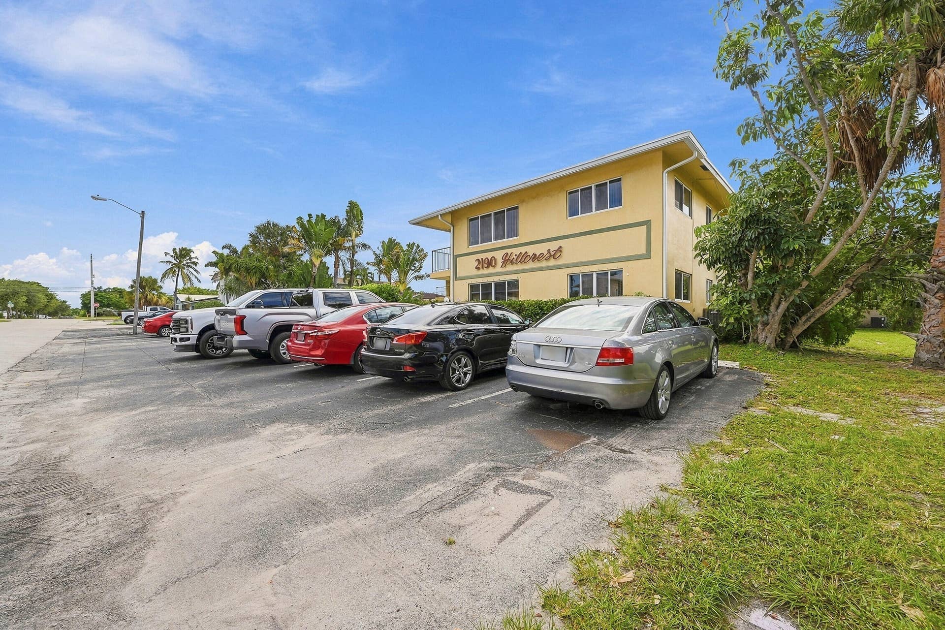 Multi-story building exterior featuring a low-pitch roofline, stucco-finish facade, designated surface parking, and mature tropical landscaping