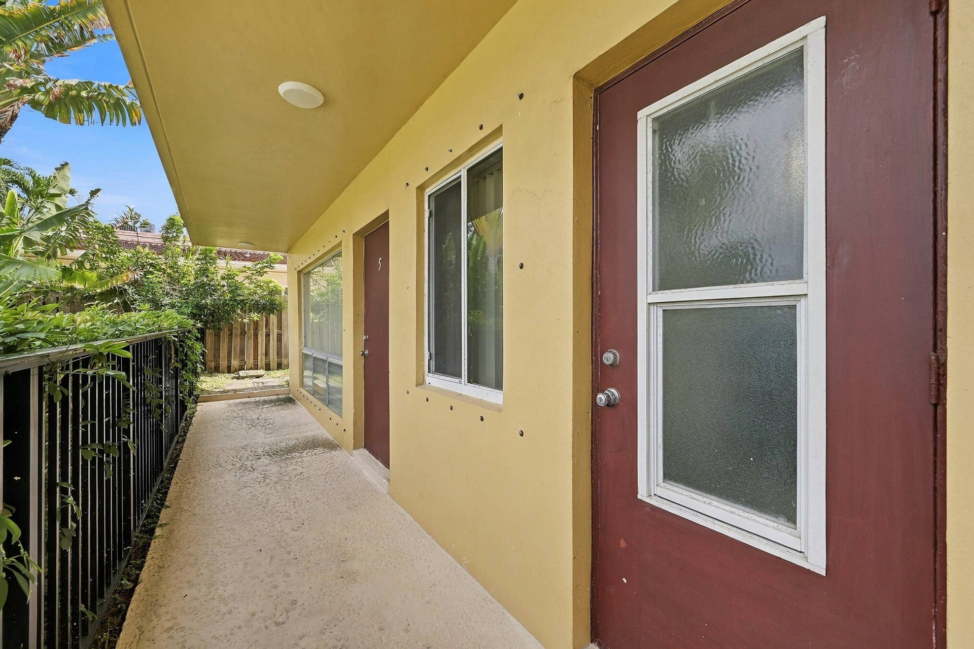 Covered exterior walkway featuring a concrete surface, sliding glass windows, doors with inset textured glass, and a metal perimeter fence