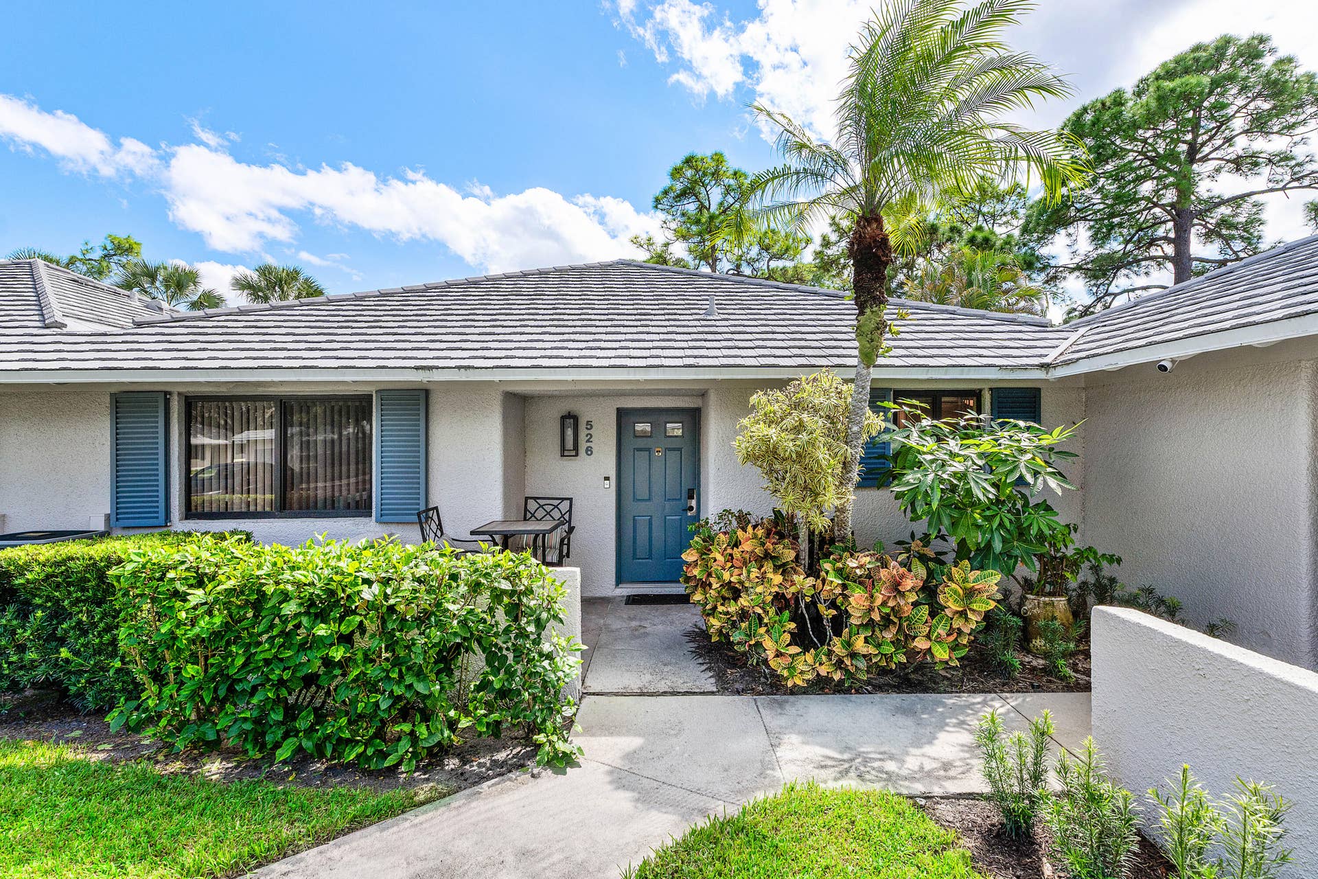 526 Club Drive, Palm Beach Gardens, FL 33418 Stucco exterior residence featuring a gray concrete tile roof, solid door with glass detailing, decorative blue shutters, and a paved walkway leading to the entryway