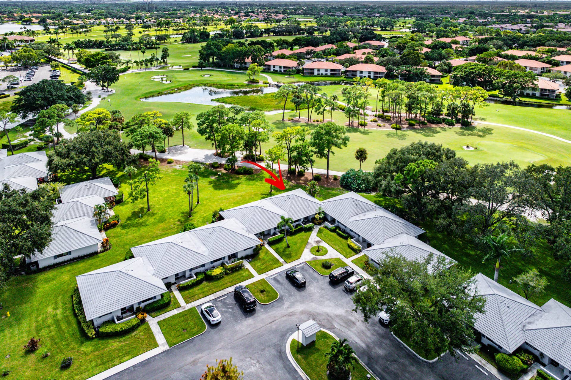 526 Club Drive, Palm Beach Gardens, FL 33418 Aerial perspective of a multi-unit complex featuring light-colored shingle roofing, paved parking areas, and mature landscaping, positioned alongside a golf course with water features and expansive green space