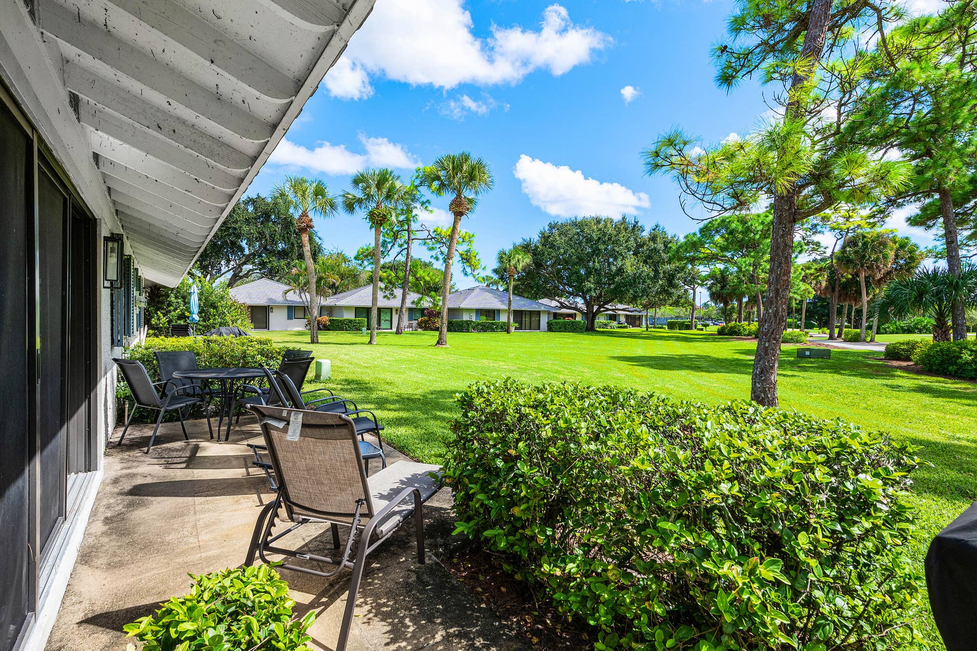 526 Club Drive, Palm Beach Gardens, FL 33418 Covered concrete patio with sliding glass door access, overlooking an expansive lawn featuring mature trees and established palm plantings