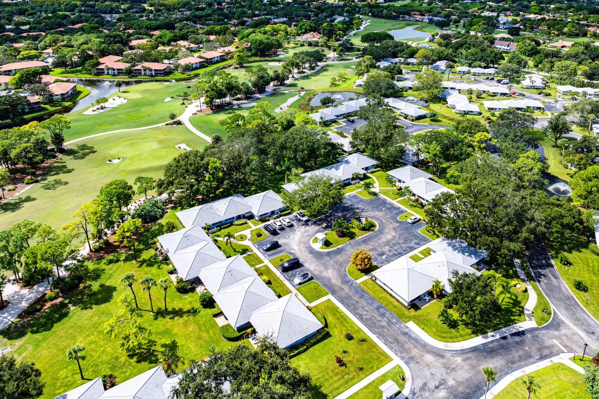 526 Club Drive, Palm Beach Gardens, FL 33418 Aerial view of a residential complex featuring light-colored hip roofs, paved parking areas, extensive green space, mature trees, and direct proximity to a golf course