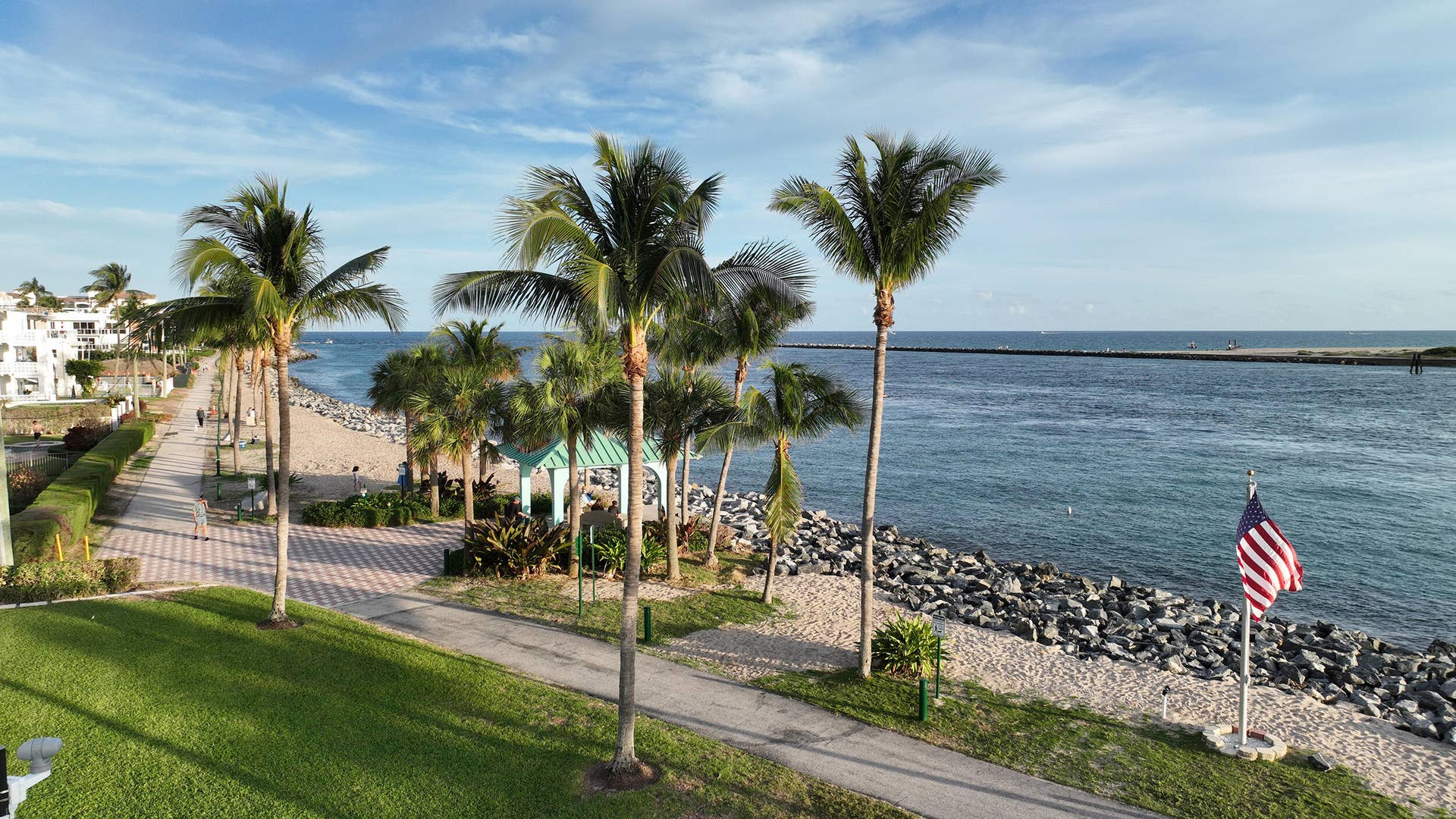 184 Lake Drive, #4201, Palm Beach Shores, FL 33404 Waterfront property featuring a paved promenade, mature palm trees, expansive lawn, and a rock-lined shoreline