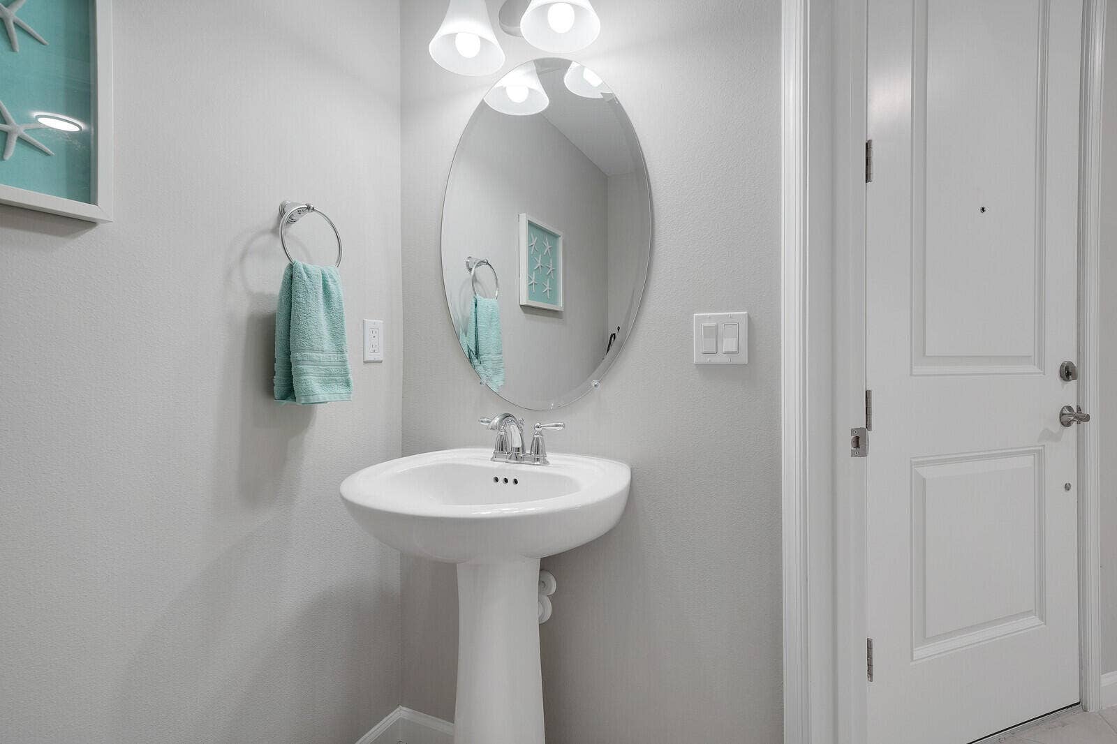 White pedestal sink with chrome faucet, oval frameless mirror, and three-light vanity fixture